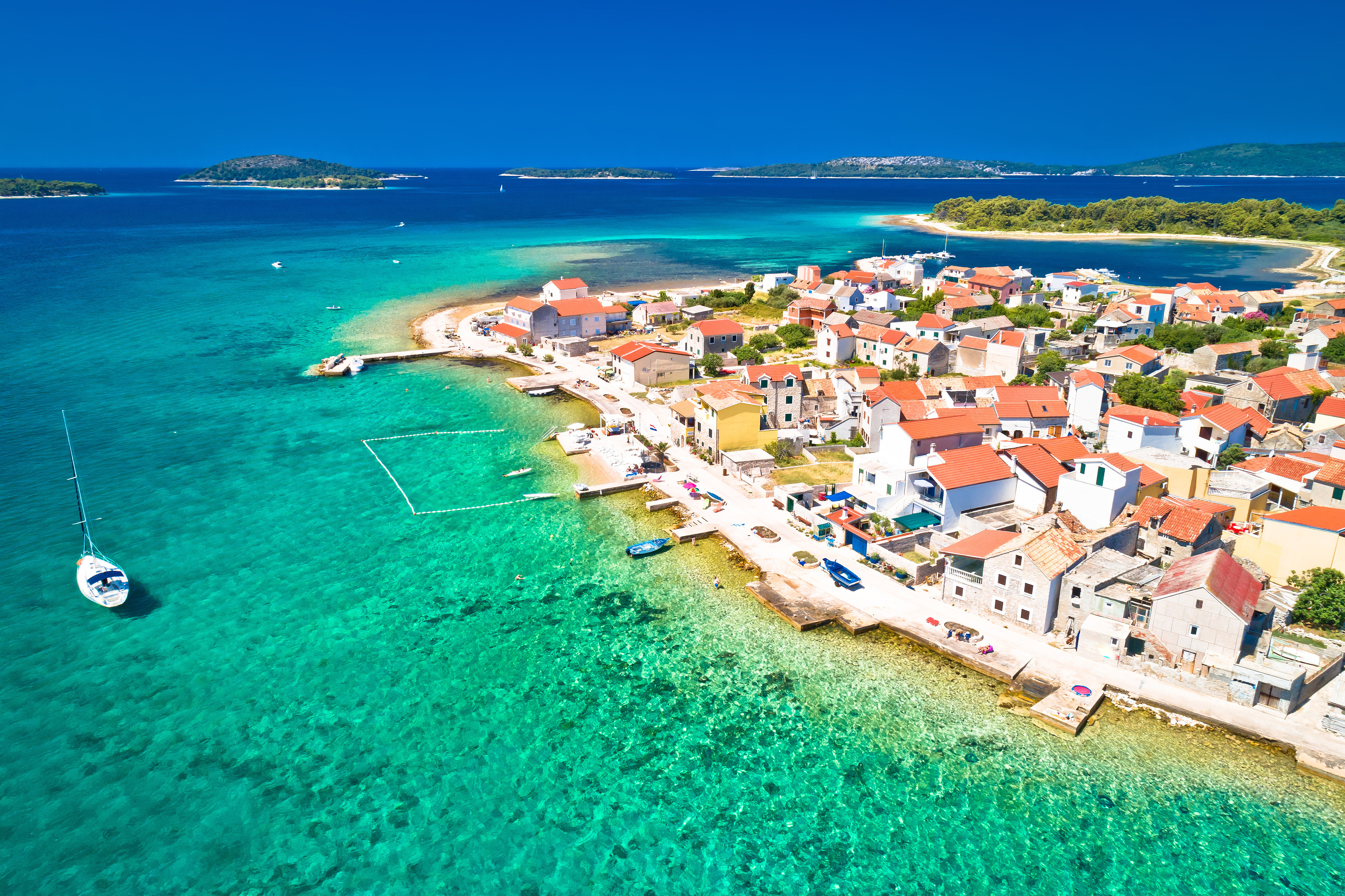 Aerial view of a coastal Mediterranean village with bright red-roofed houses, turquoise sea, and a docked sailboat, surrounded by lush greenery and islands on a clear day