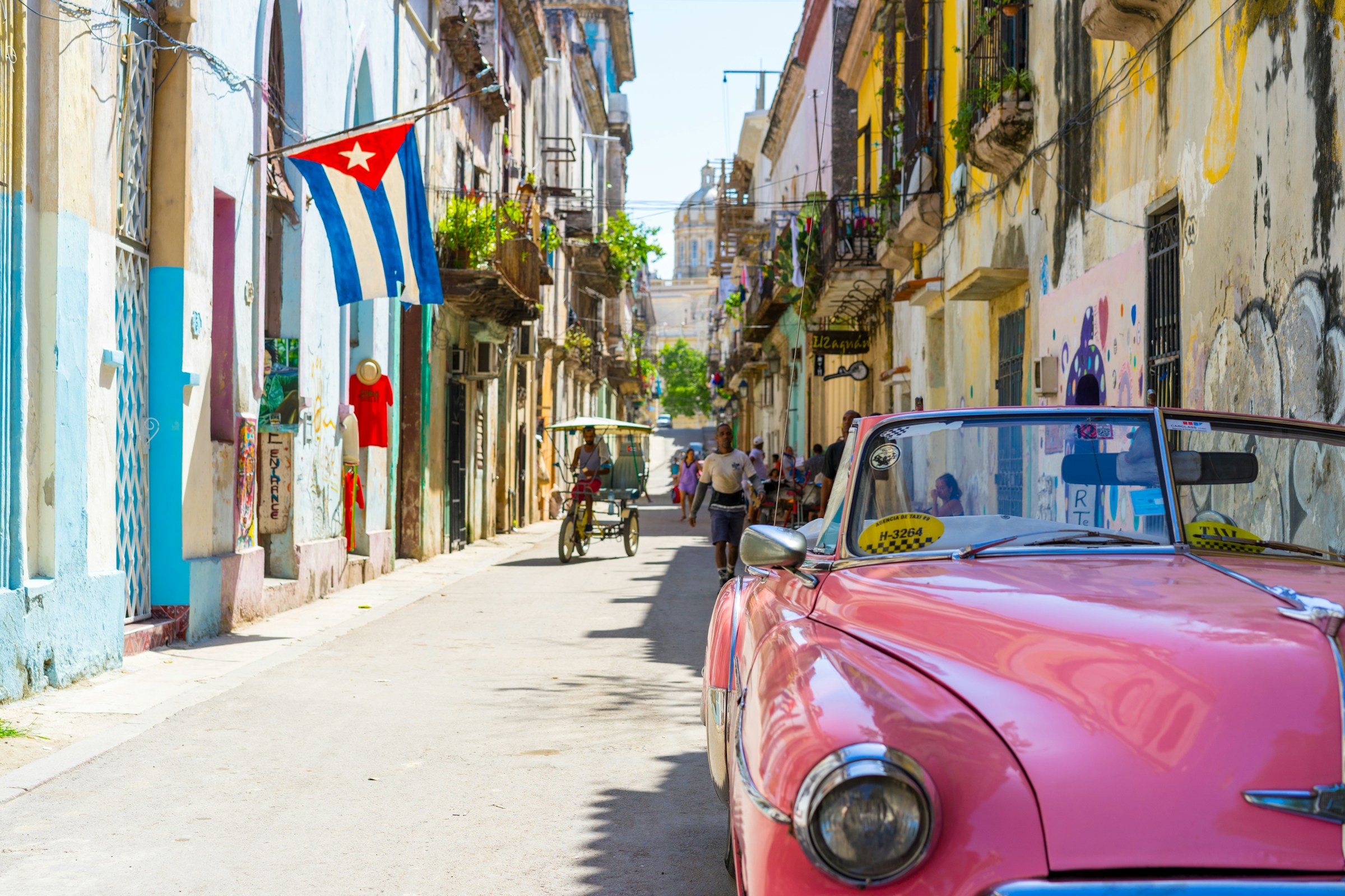 Vintage pink convertible car parked on a narrow, colorful street in Havana, Cuba, with a Cuban flag displayed on a nearby building and people walking and biking in the background