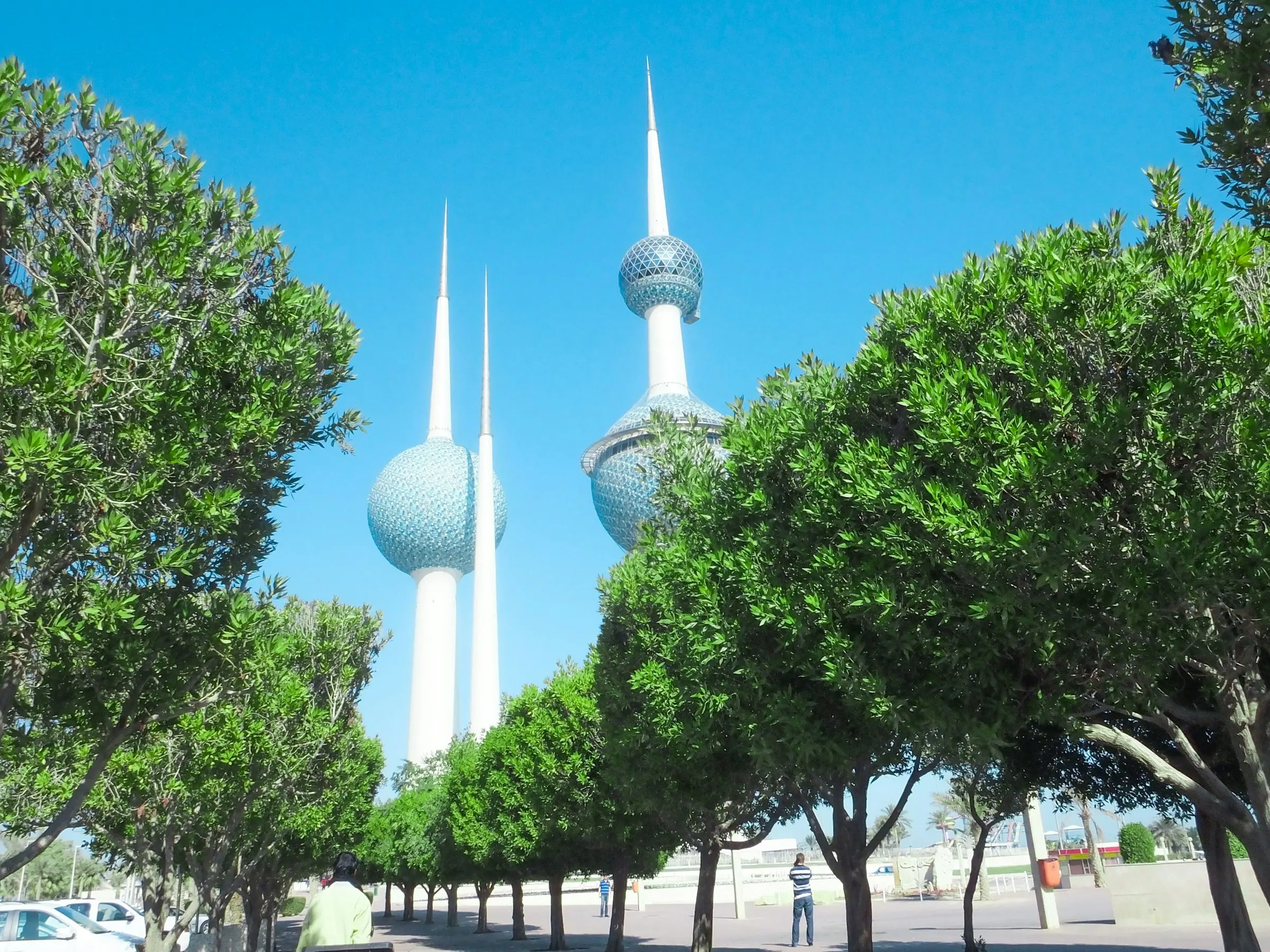 Kuwait Towers against clear blue sky, surrounded by lush green trees in a park setting.