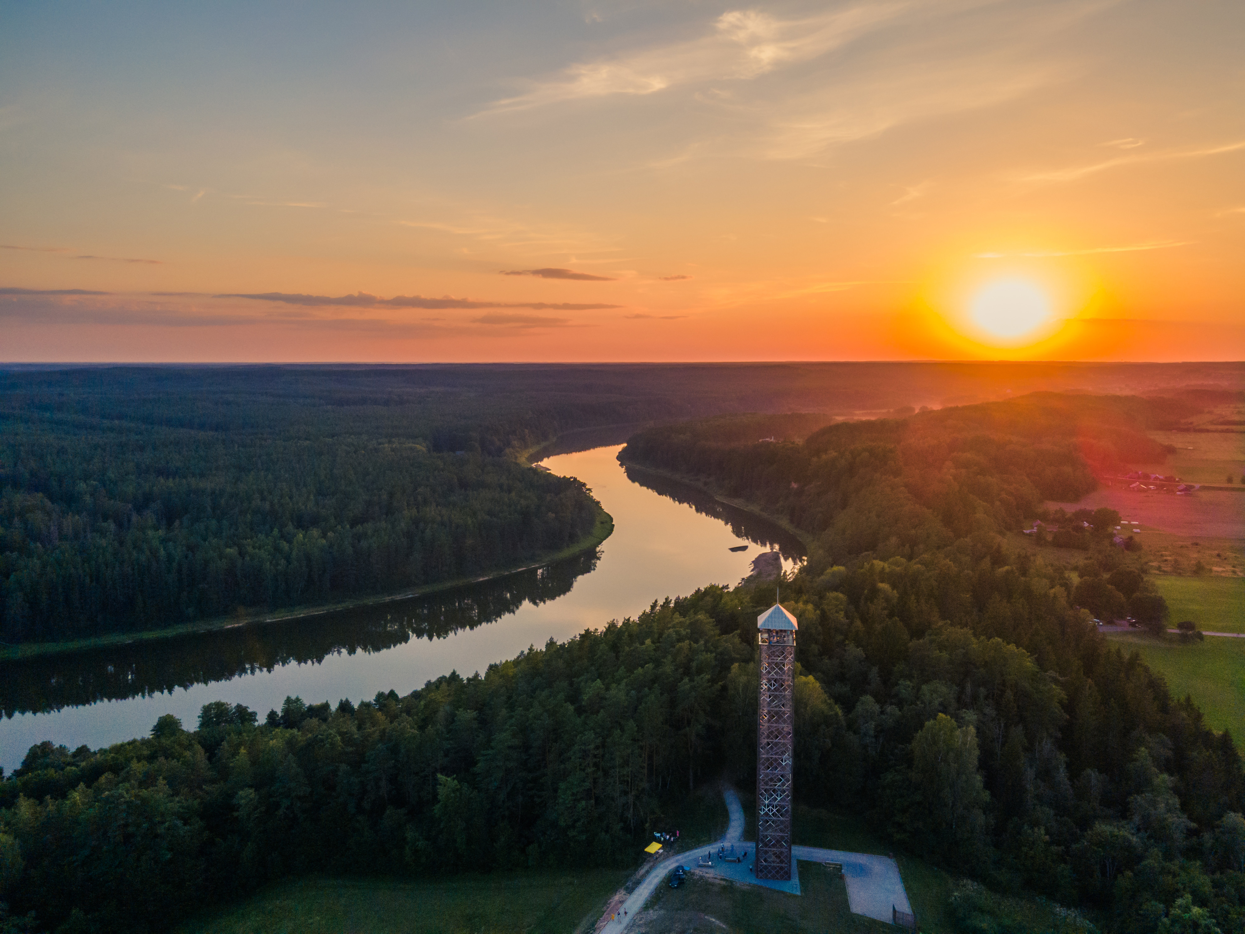 Sunset over a scenic river with lush forests and a tall observation tower, offering panoramic views of Lithuania.