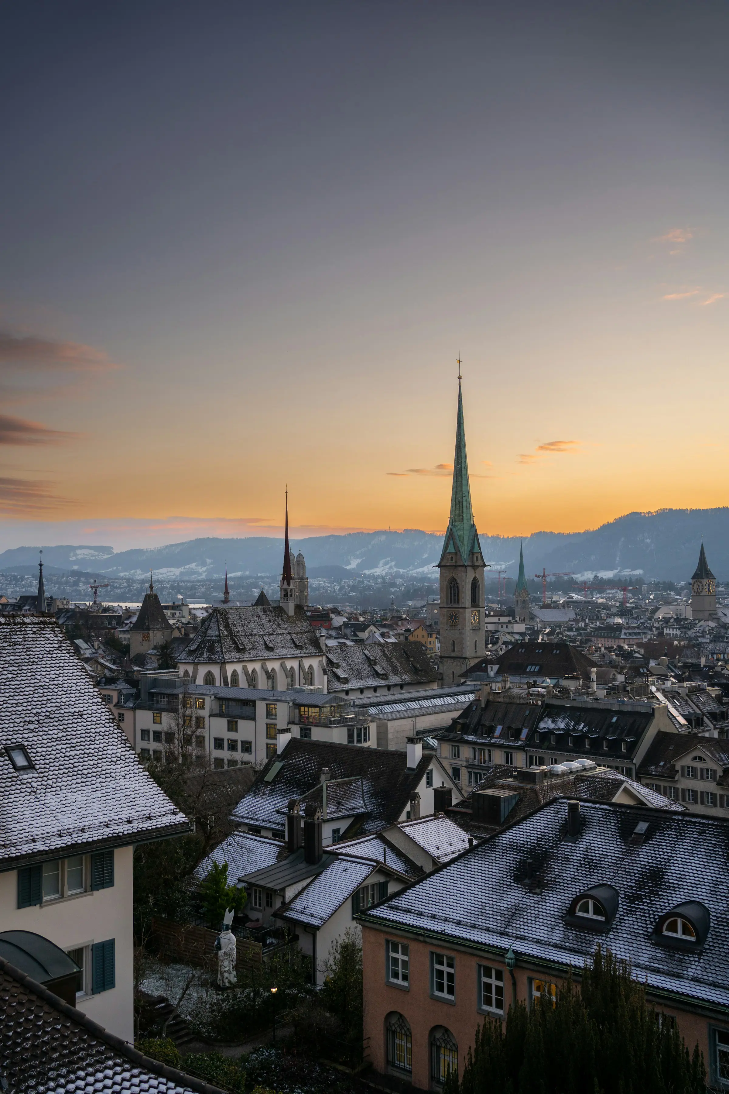Scenic view of Zurich, Switzerland at sunset with snow-dusted rooftops and church spires against a mountainous backdrop