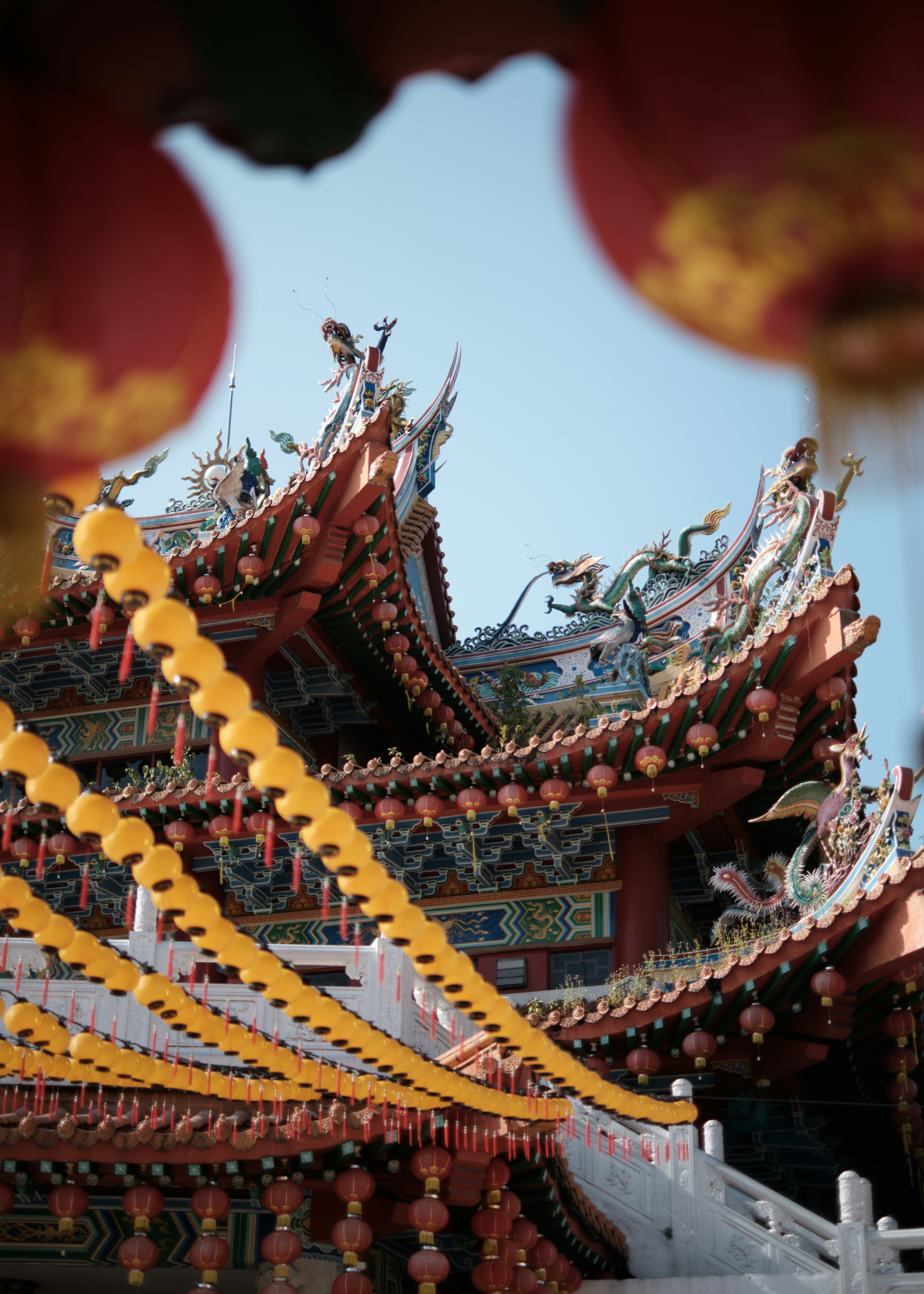 Traditional Chinese temple with ornate curved rooftops and decorative dragon sculptures, framed by vibrant yellow and red lanterns, set against a clear blue sky