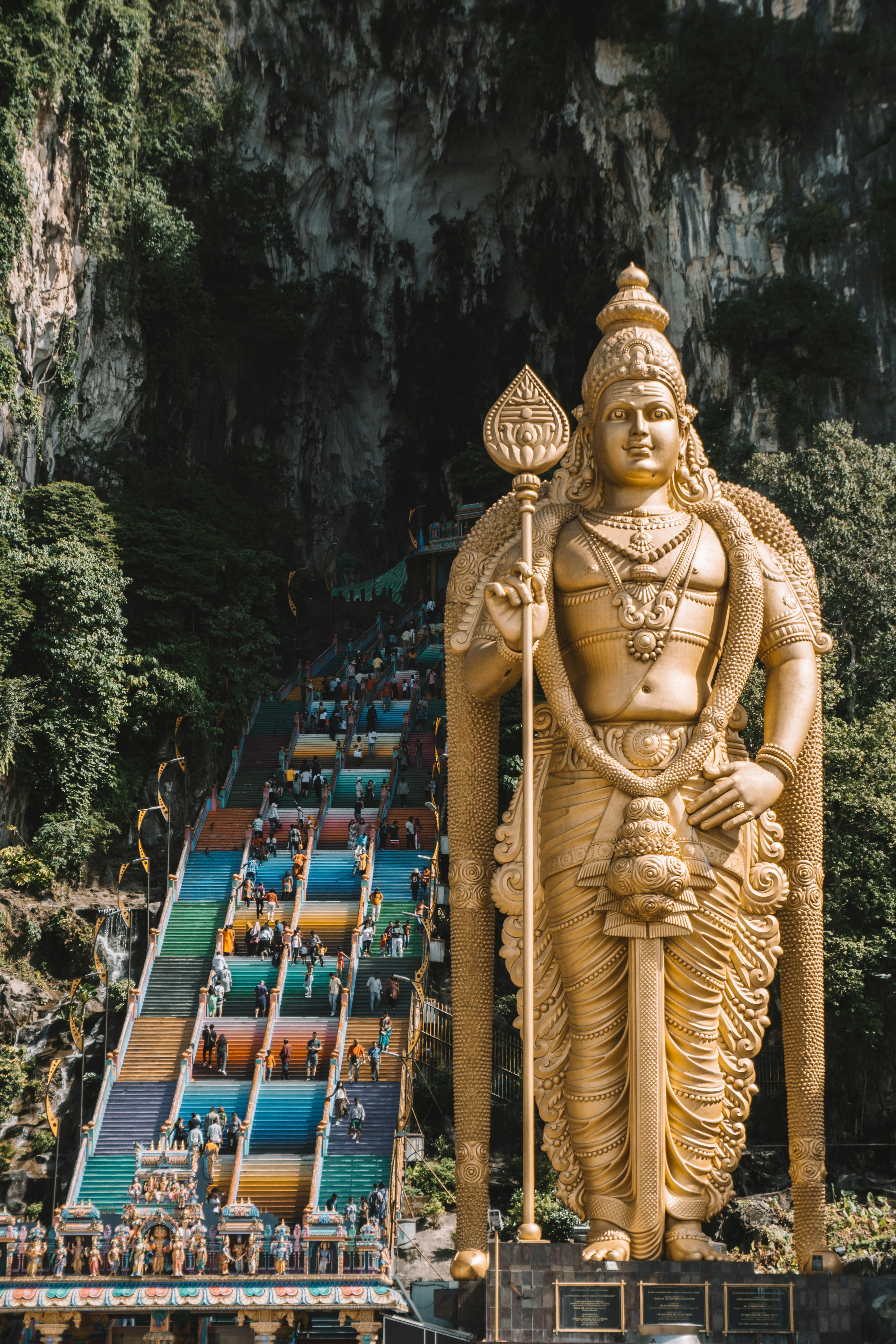 Golden statue of Lord Murugan at the entrance of Batu Caves, Malaysia, with colorful staircase leading to the cave temple, surrounded by lush green cliffs