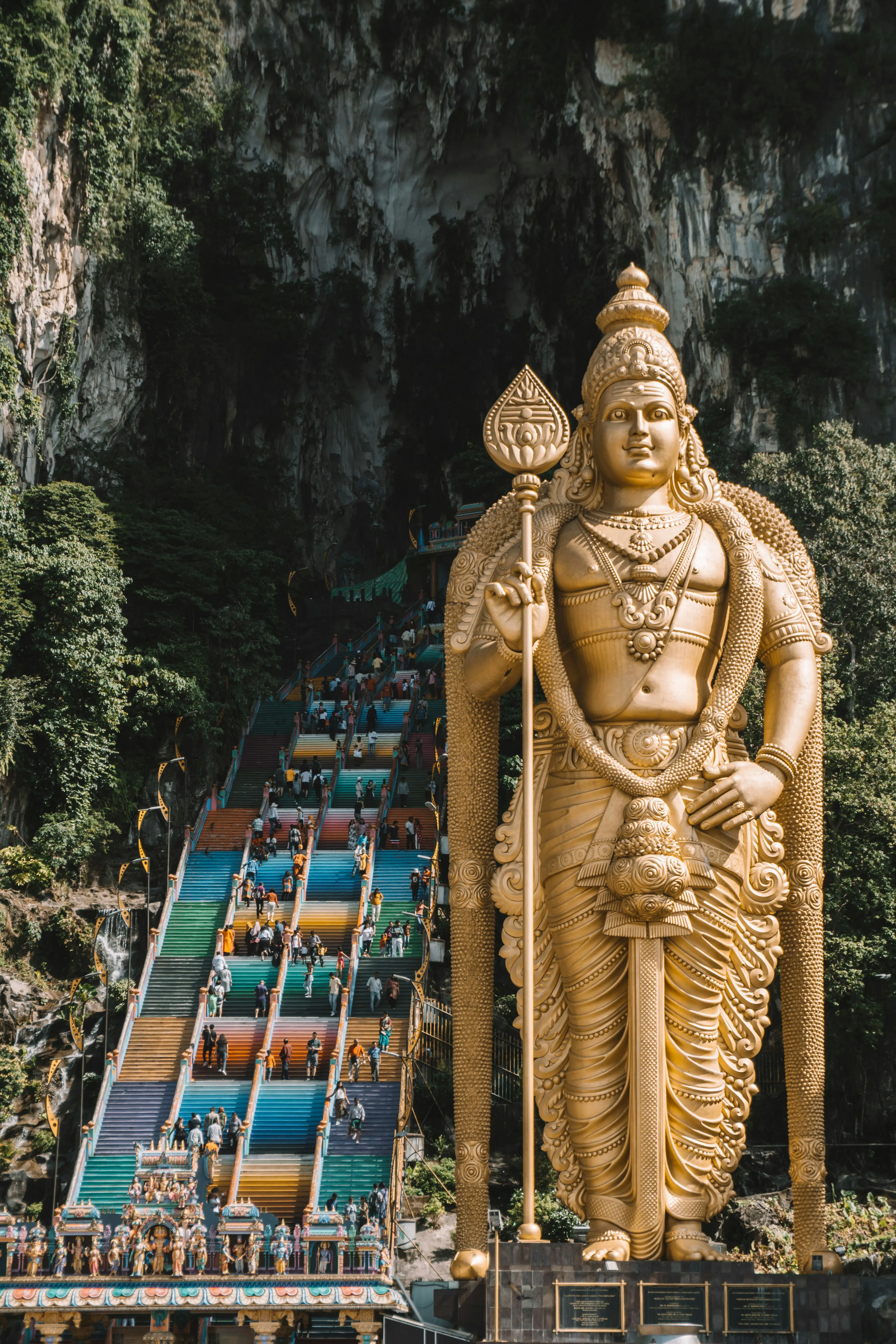 Golden statue of Lord Murugan at the entrance of Batu Caves, Malaysia, with colorful staircase leading to the cave temple, surrounded by lush green cliffs