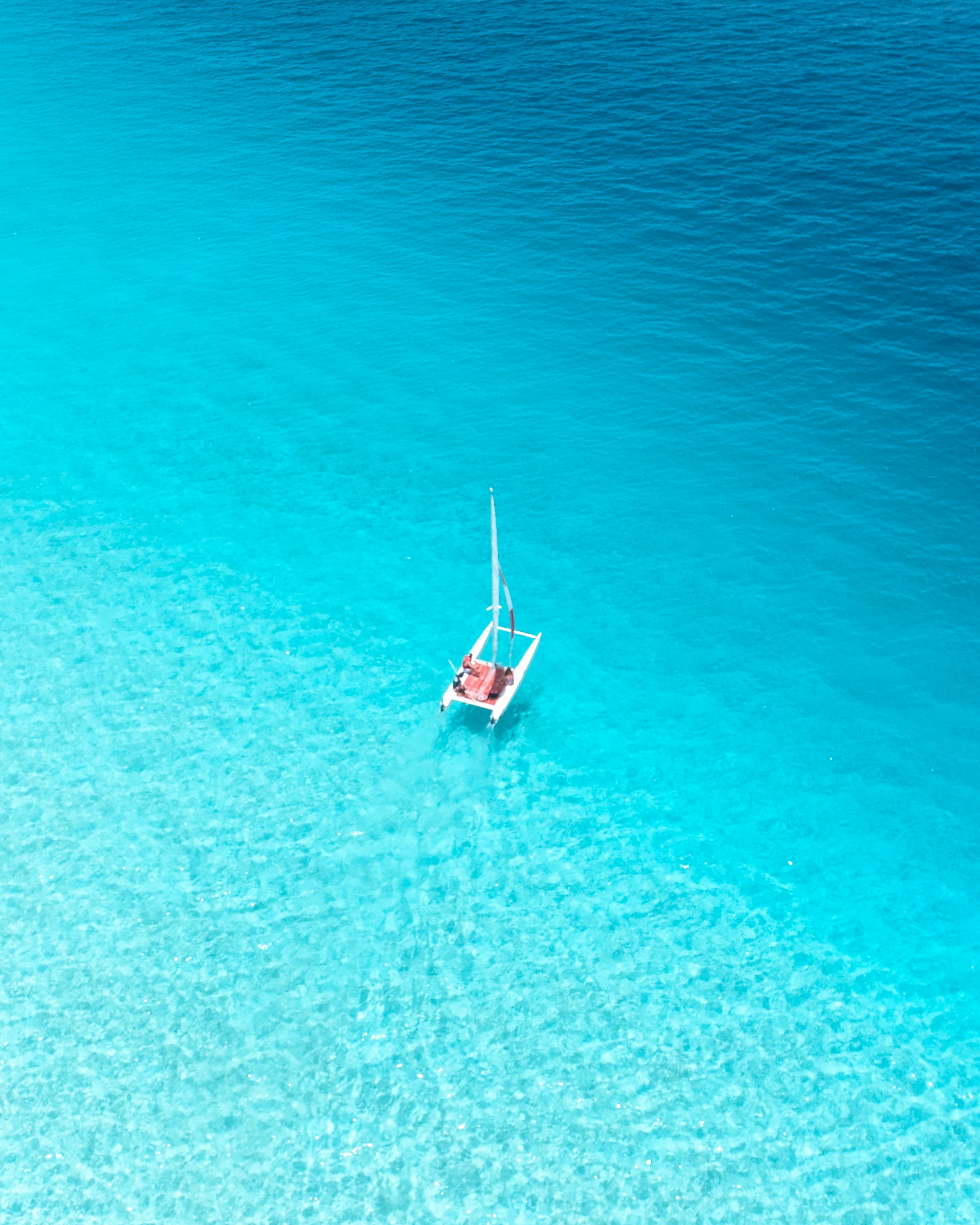 Aerial view of a small sailboat on a vast expanse of clear turquoise water, highlighting the serene and tropical ocean setting