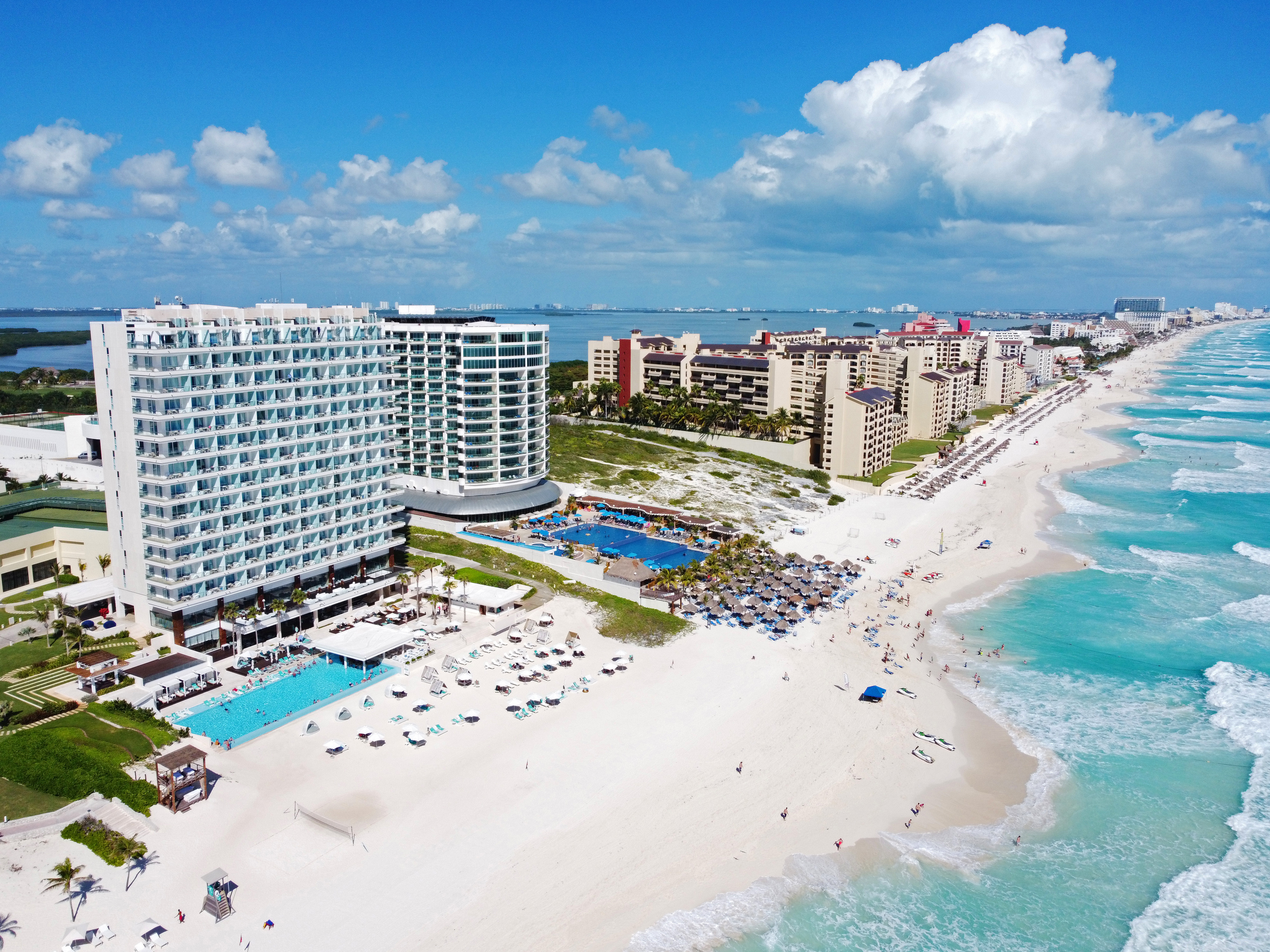 Aerial view of a beach resort in Cancún, Mexico, featuring sandy beaches, turquoise ocean waves, and modern hotel buildings under a partly cloudy sky