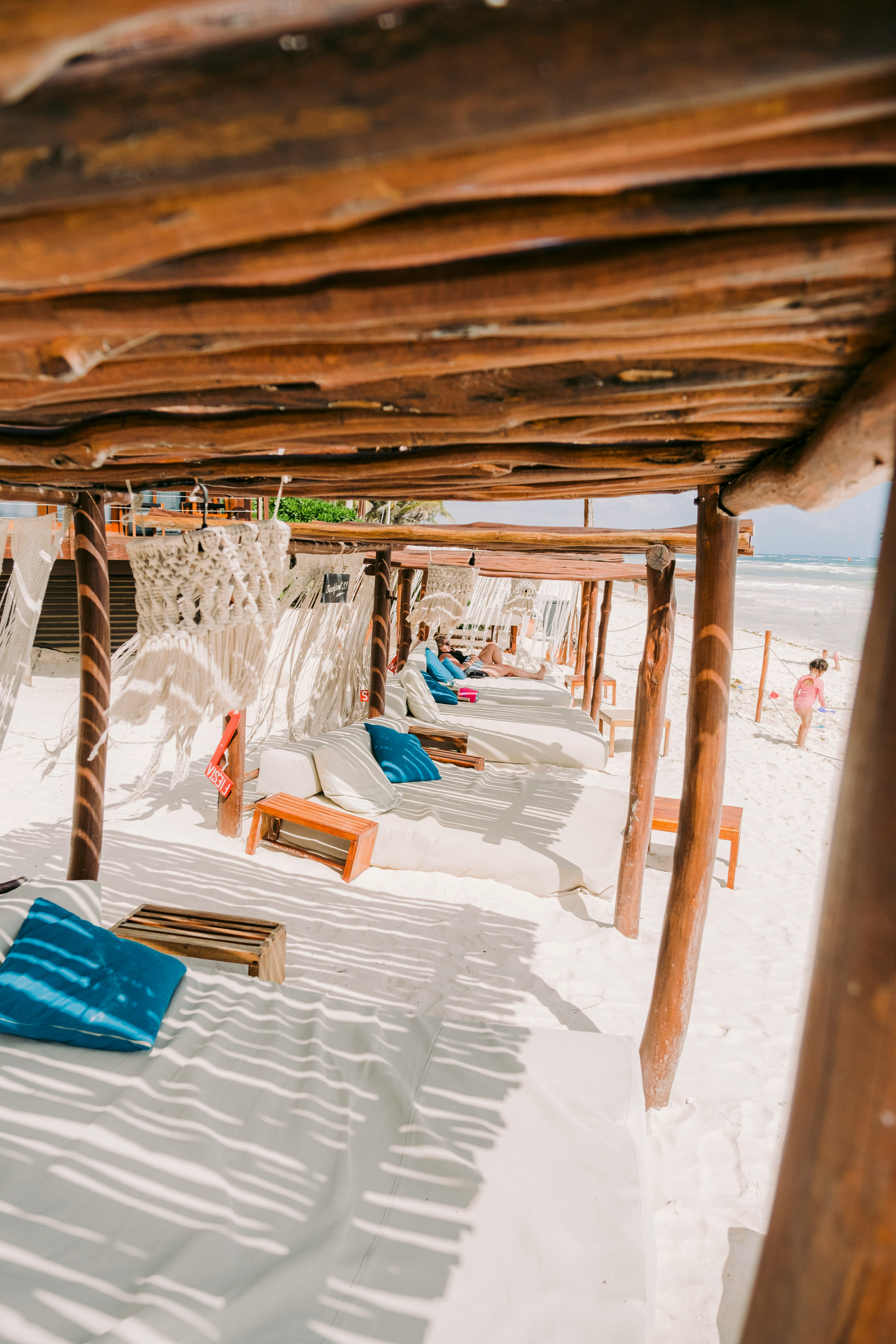 Beach cabanas with white sand and blue cushions under wooden pergolas, offering a relaxing seaside experience