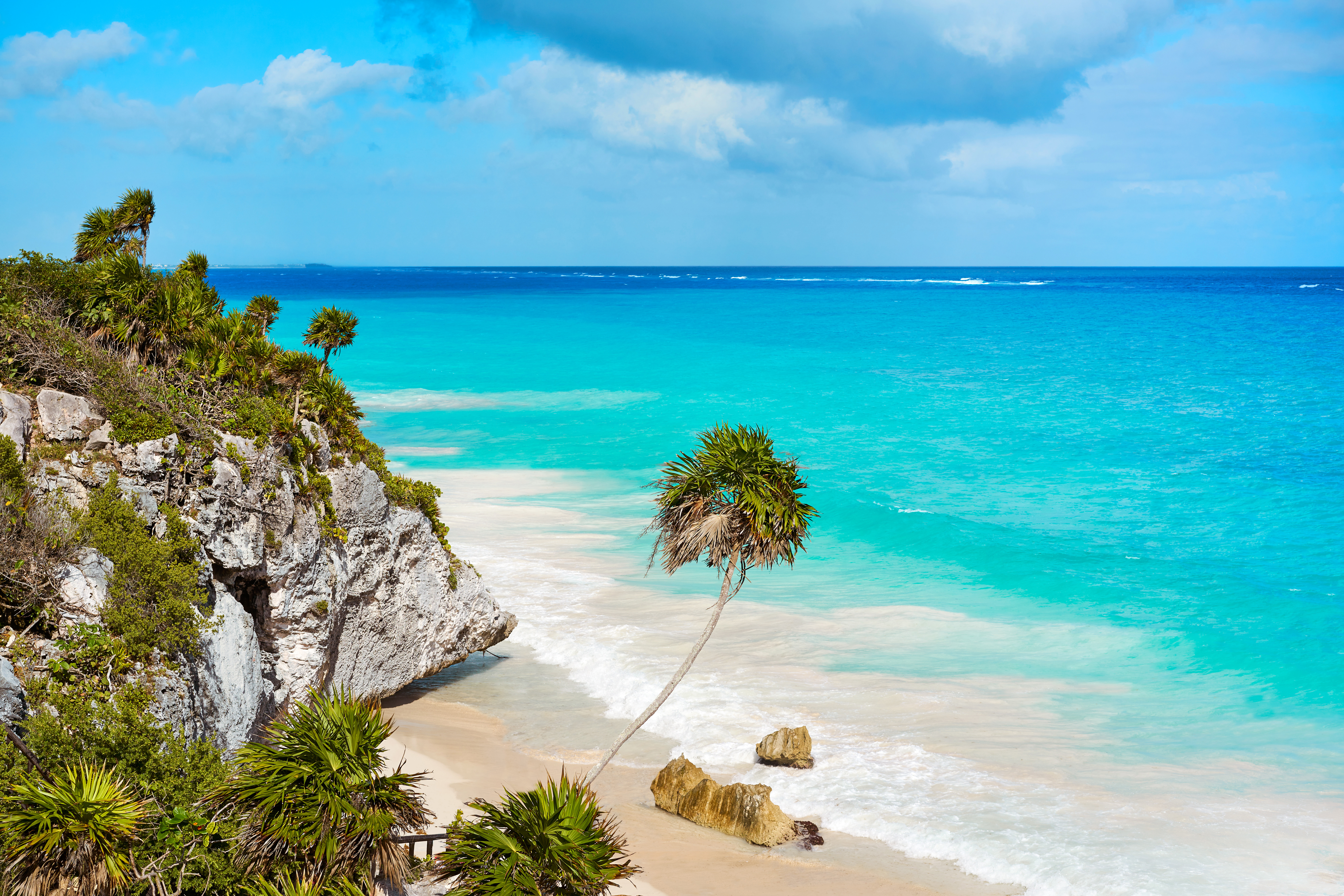 Scenic view of Tulum beach with turquoise waters, rocky cliffs, and palm trees under a partly cloudy sky