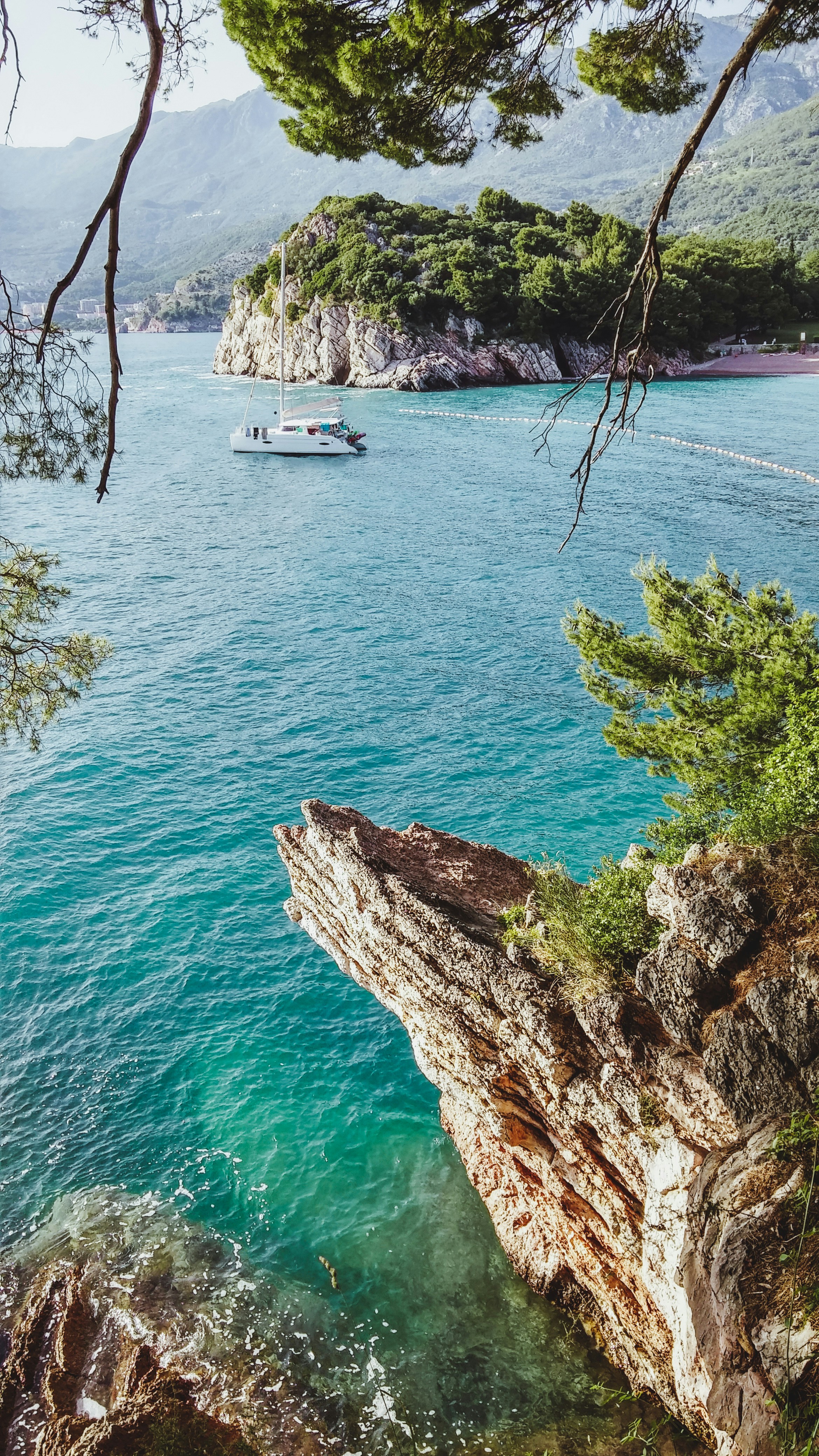 Scenic coastal view with a sailboat on turquoise waters near a rocky cliff and lush green island under a clear sky