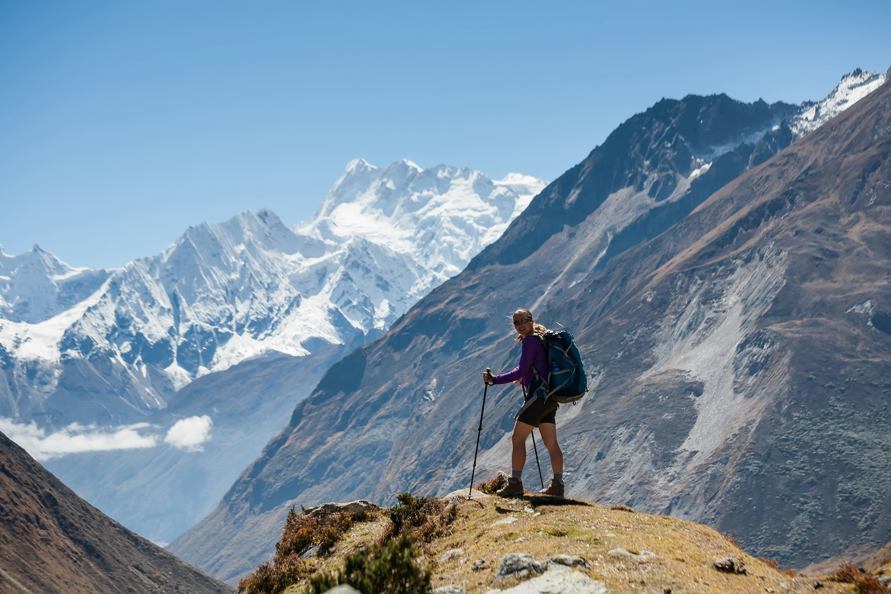 Hiker with trekking poles navigating a mountain trail with snow-capped peaks in the background under a clear blue sky