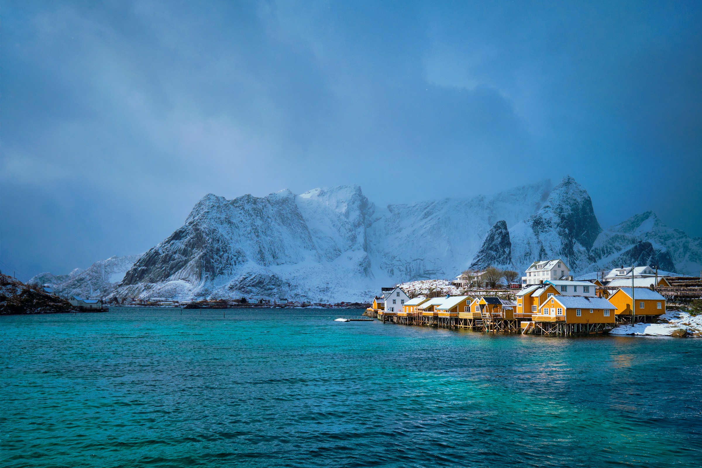 Snow-covered mountains and turquoise water surround traditional yellow cabins on stilts in Lofoten, Norway.