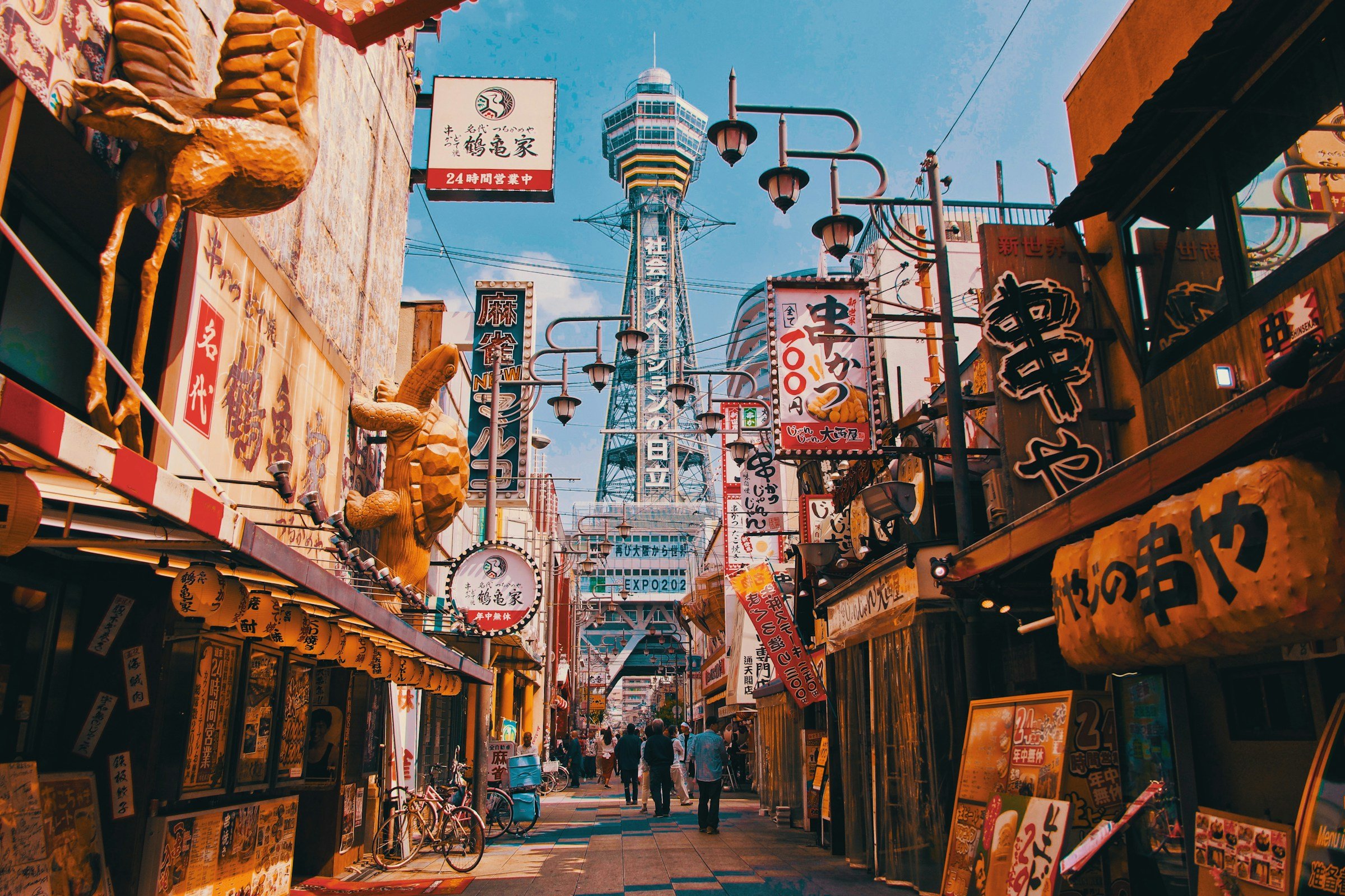 Livlig gata i Osaka, Japan, med restauranger, skyltar och utsikt mot Tsutenkaku-tornet under klarblå himmel.