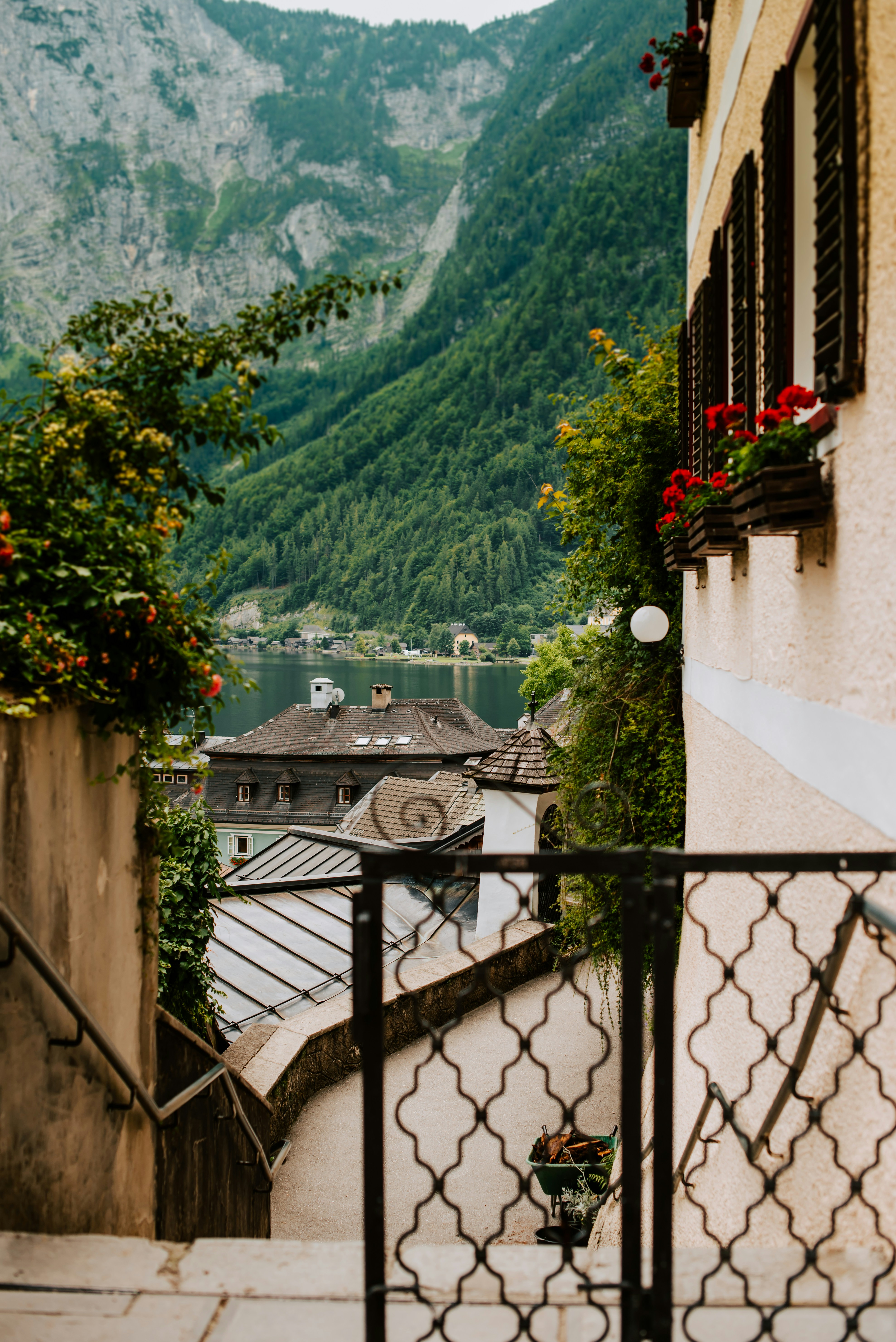 Scenic view of a European village street with ornate railing, overlooking rooftops and a lush green mountain landscape. Red flowers adorn a yellow house, adding vibrant color