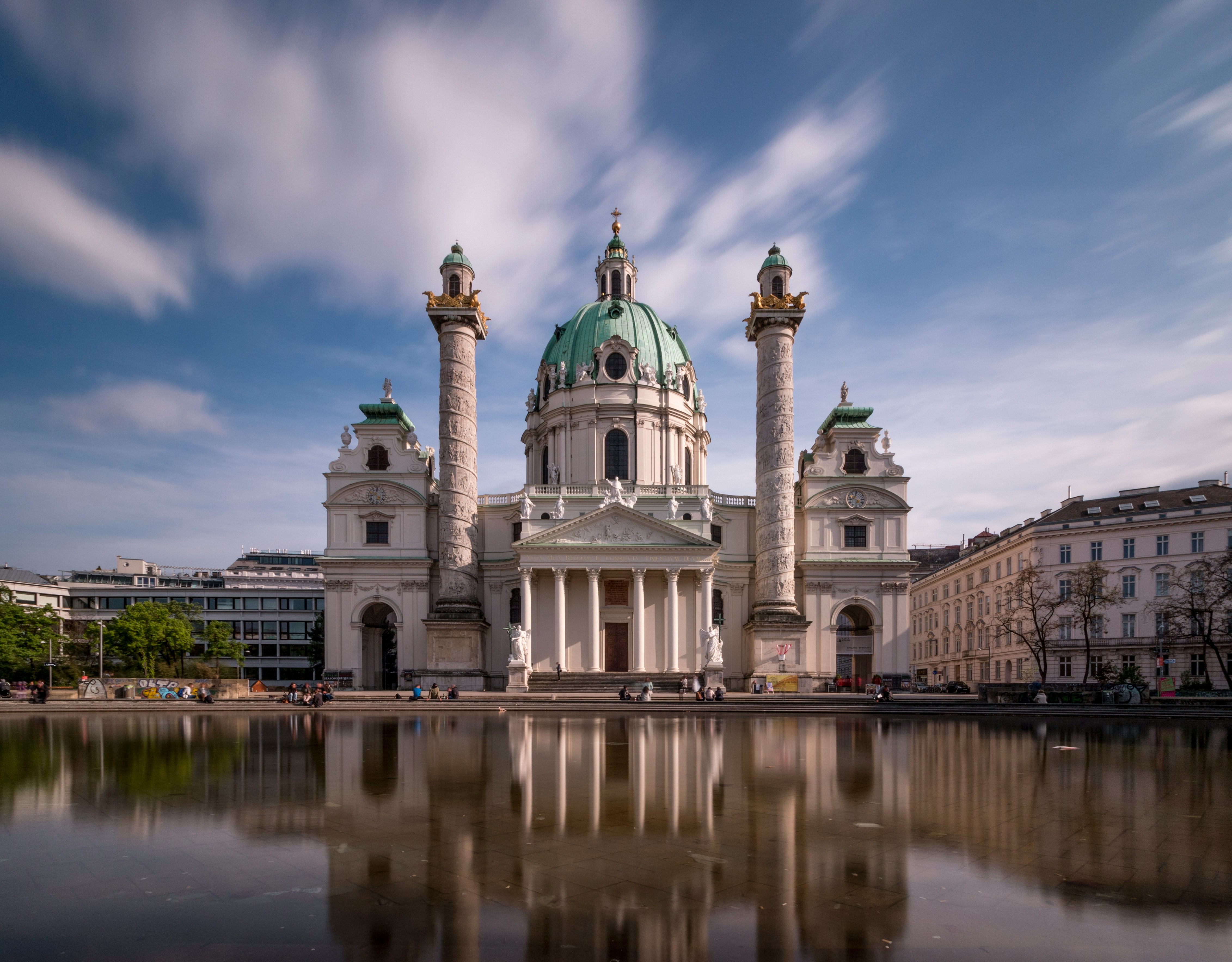 St. Charles Church in Vienna, Austria with its baroque architecture and distinctive green dome, reflected in a tranquil pool under a blue sky