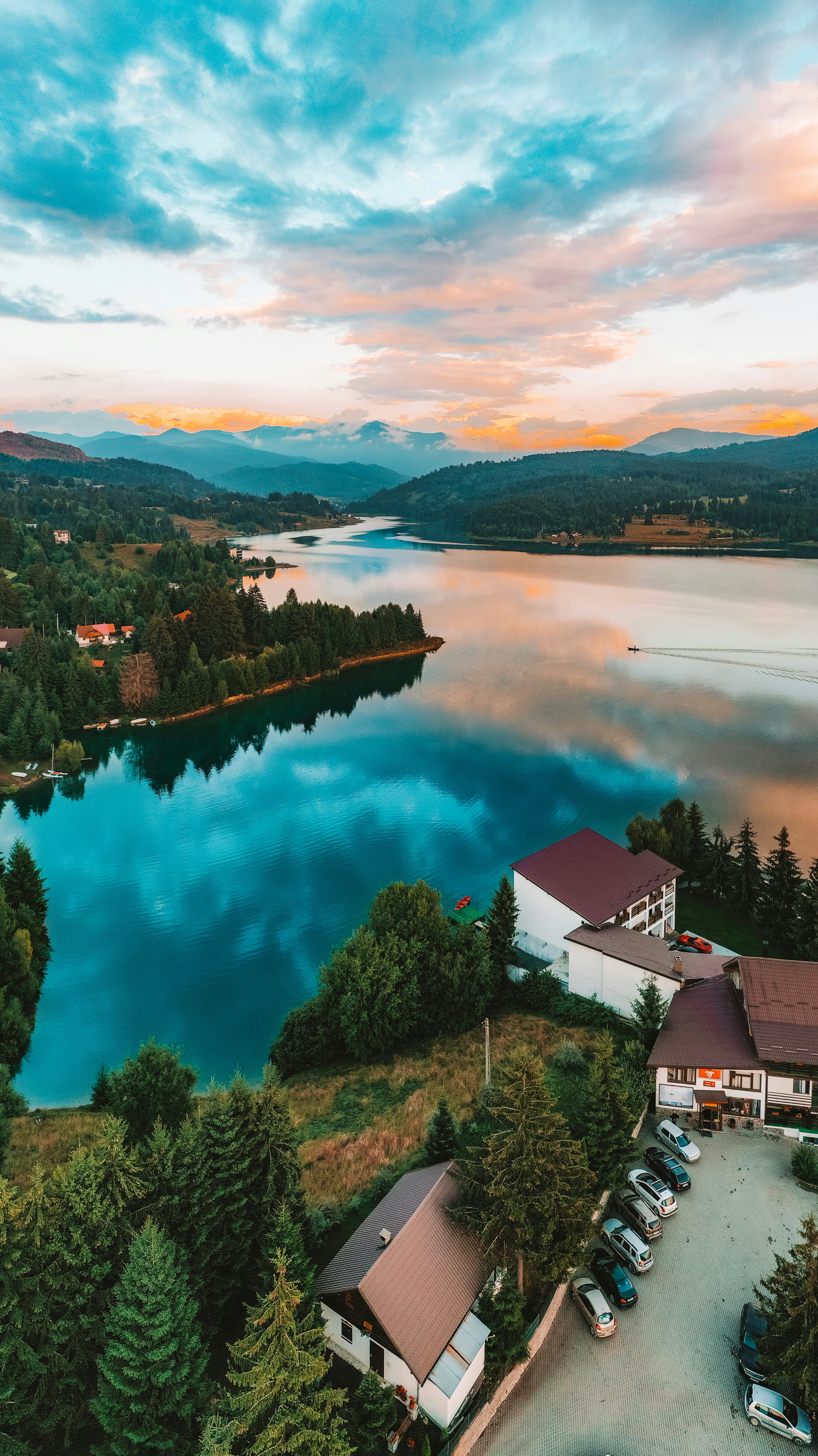 Aerial view of a serene lake reflecting a colorful sunset, surrounded by lush forests and residential buildings, with distant mountains providing a picturesque background