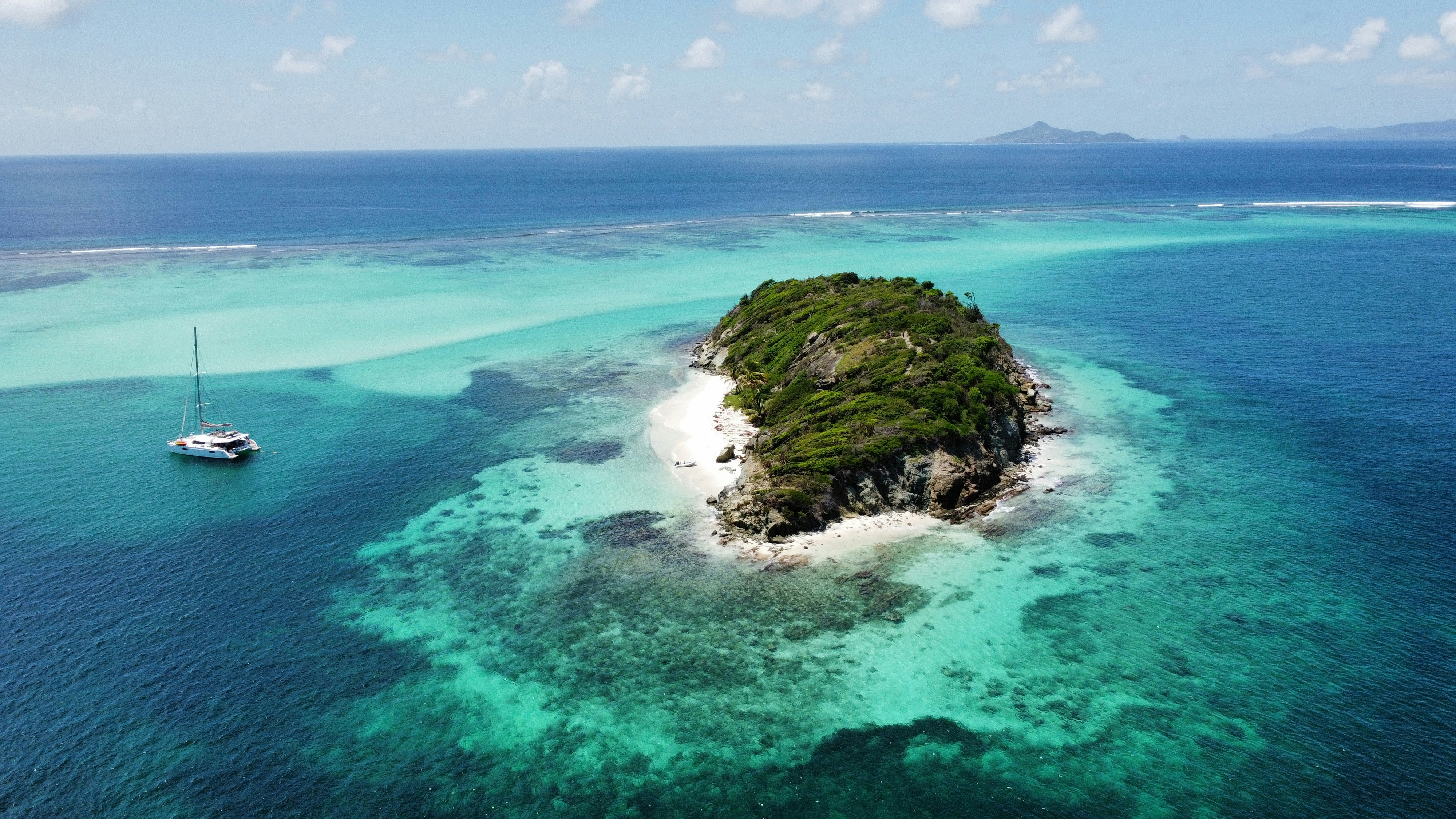 Aerial view of a picturesque tropical island with lush greenery surrounded by turquoise waters, featuring a sailboat anchored nearby