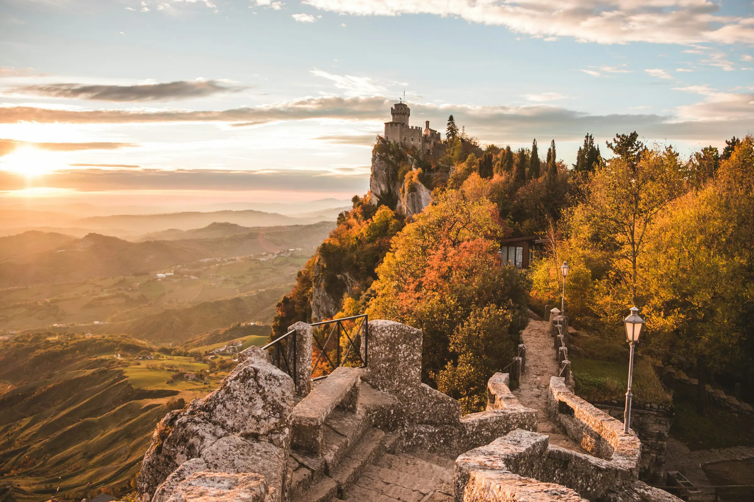 Solnedgång över ett medeltida slott på en klippa omgiven av höstfärger i San Marino, med bergig vy i bakgrunden.