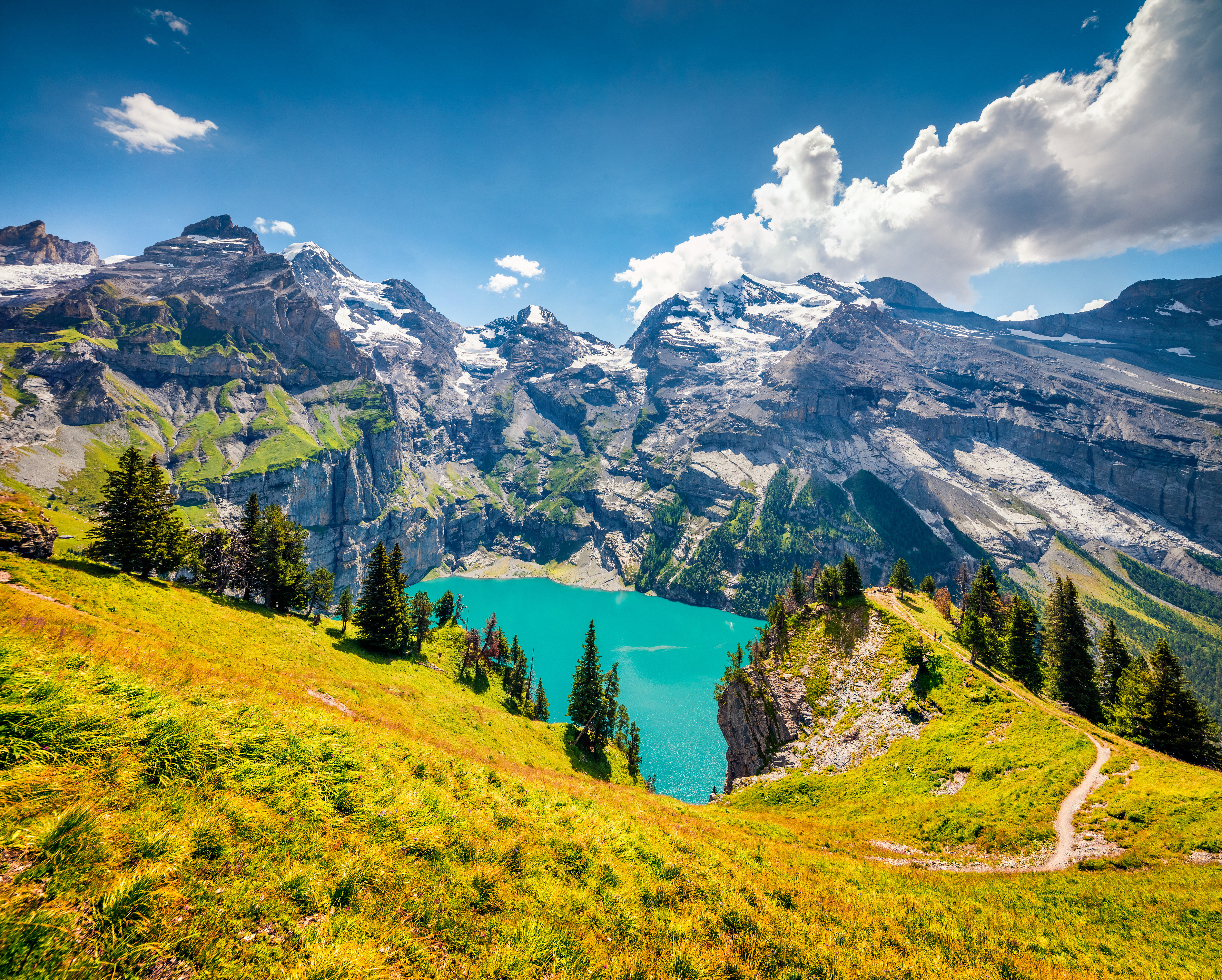 Scenic view of turquoise alpine lake surrounded by snow-capped mountains and lush greenery under a bright blue sky in the Swiss Alps