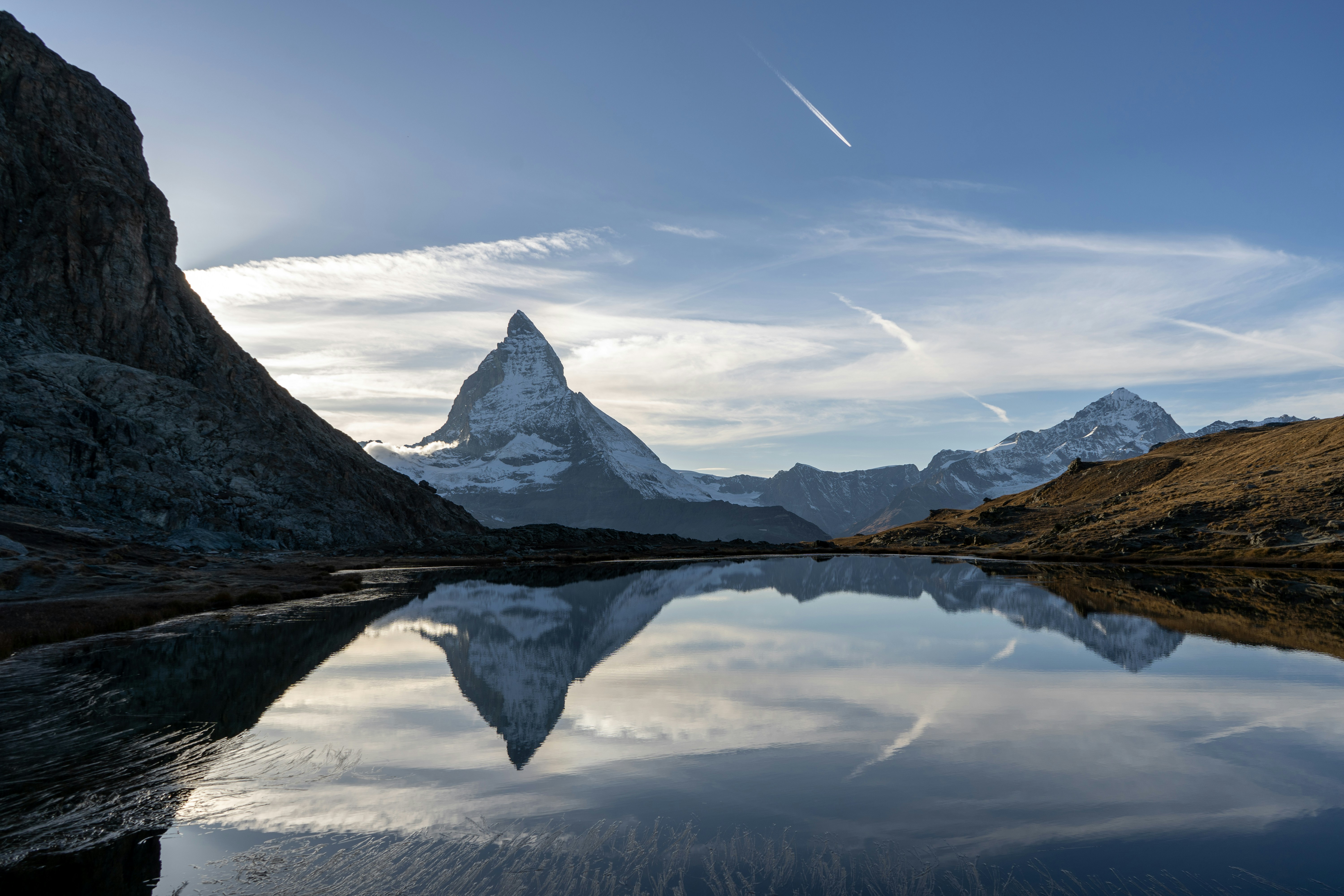 Snow-capped Matterhorn mountain reflected in a tranquil lake under a clear blue sky with clouds, surrounded by rugged terrain