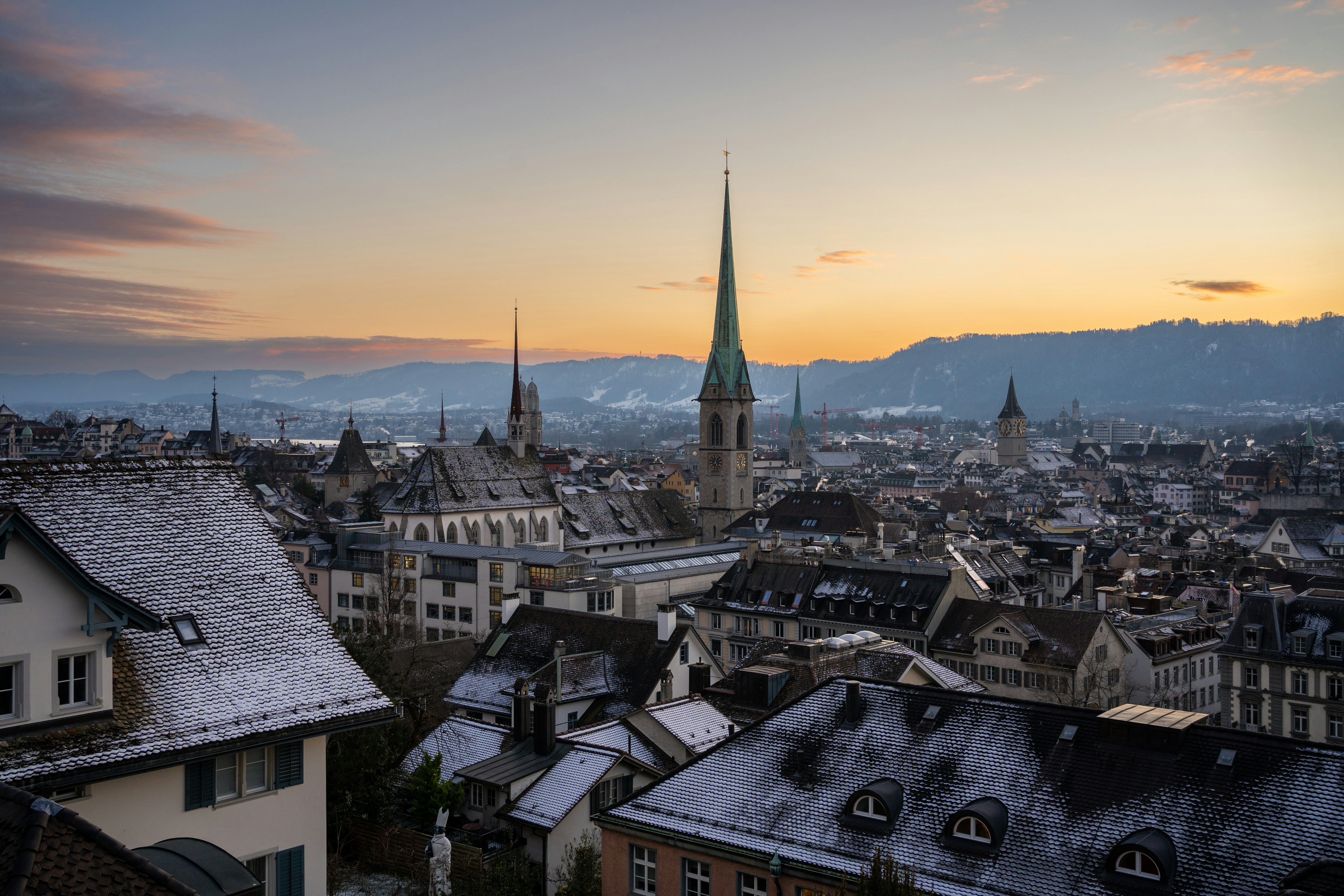 Sunrise over Zurich cityscape with snow-dusted rooftops and church spires, surrounded by distant mountains