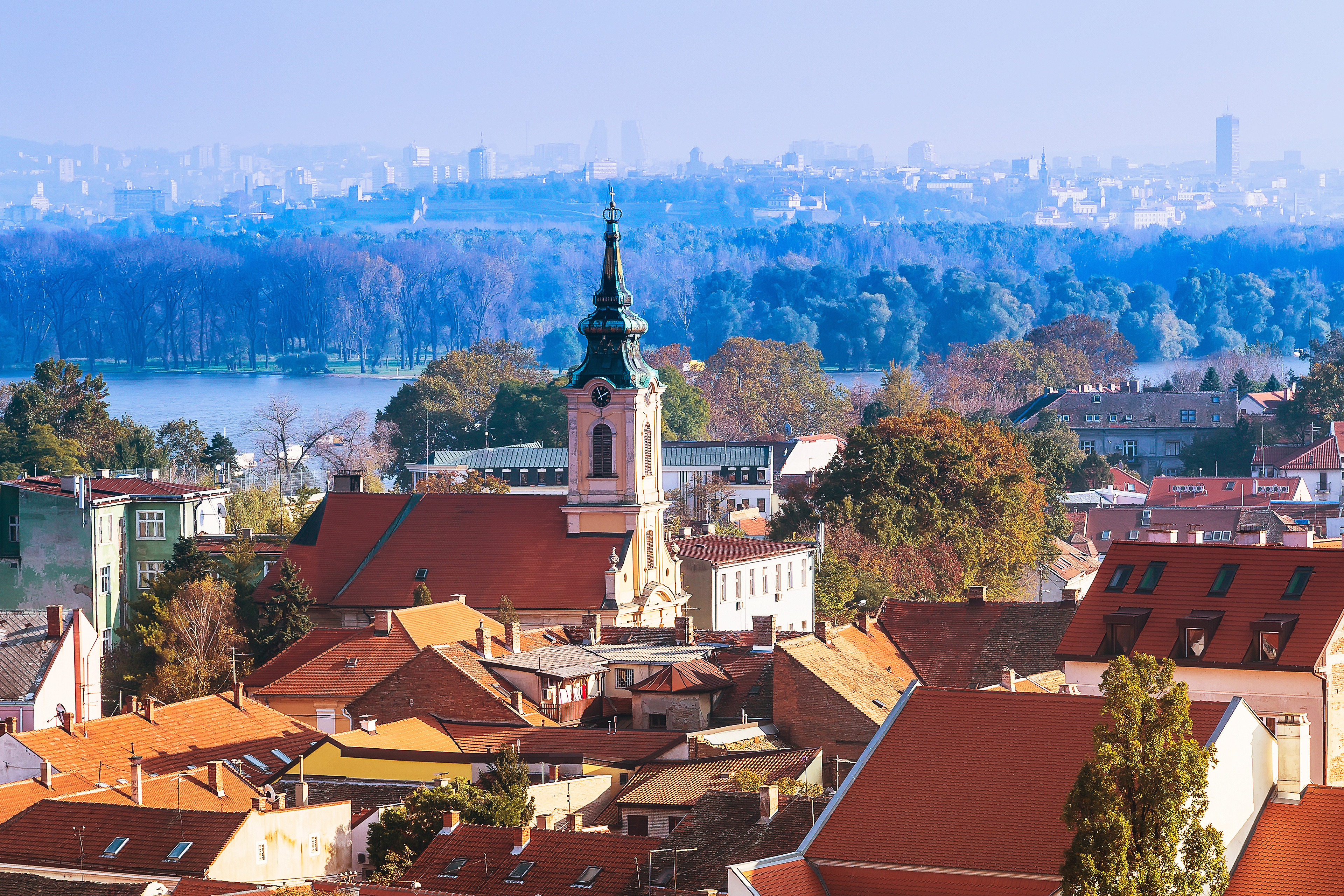 Travel to Serbia - featuring red-tiled rooftops, a historic church tower, lush trees, and the Danube River in the background.