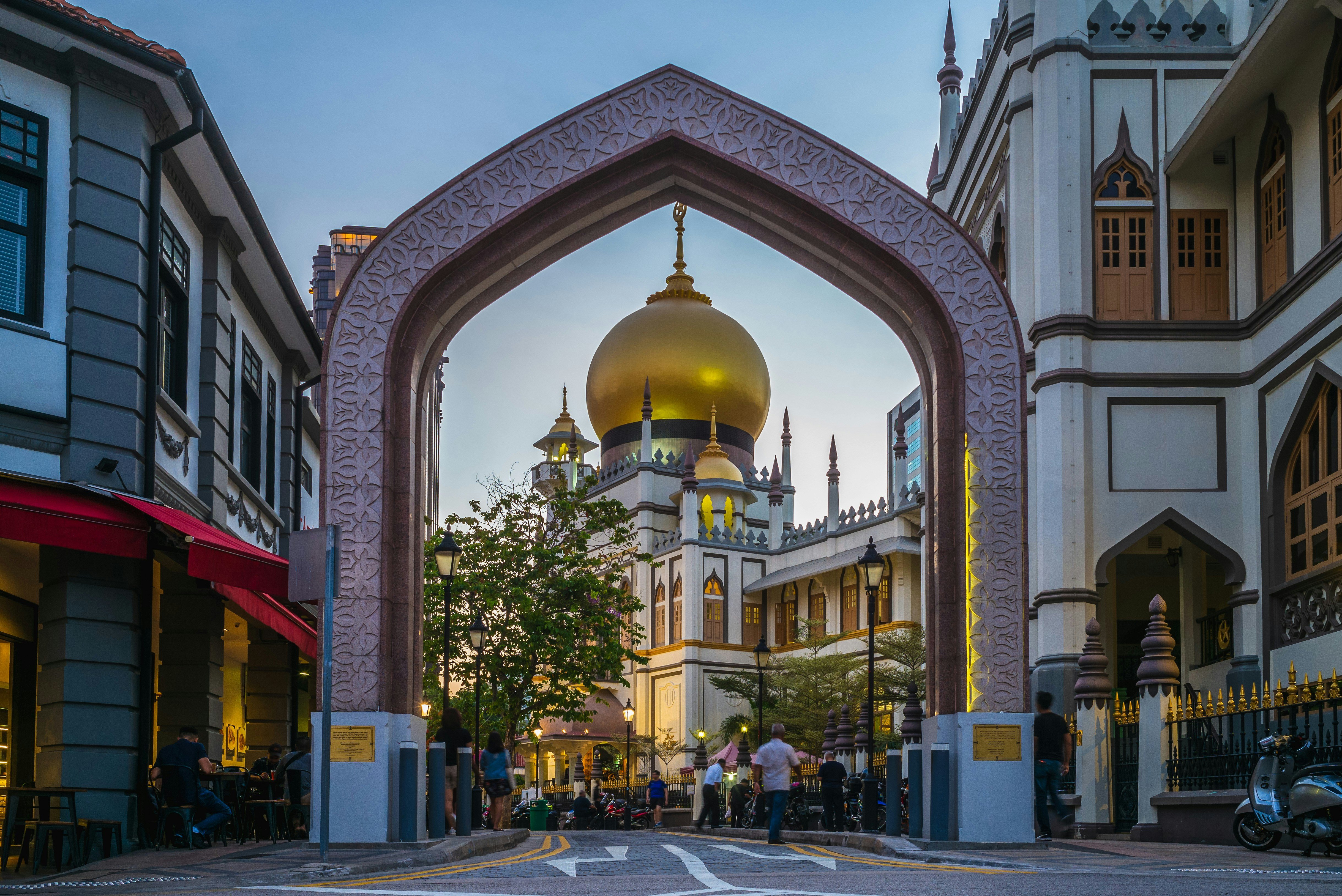 Sultan Mosque in Singapore at dusk, framed by an ornate archway, surrounded by shops and pedestrians