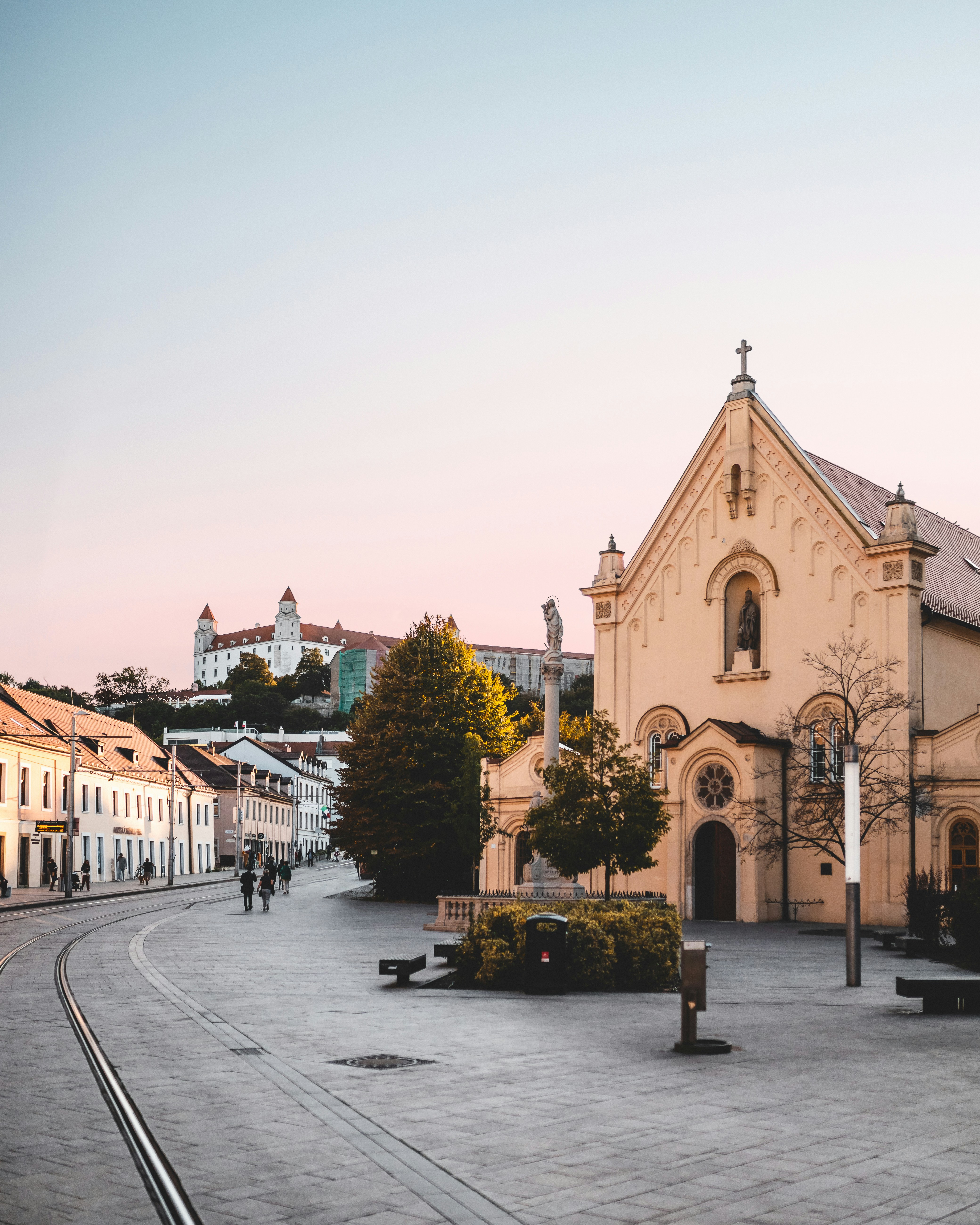 Historic European cityscape featuring a charming church and distant castle on a pastel-colored evening, with quaint buildings lining the cobblestone street