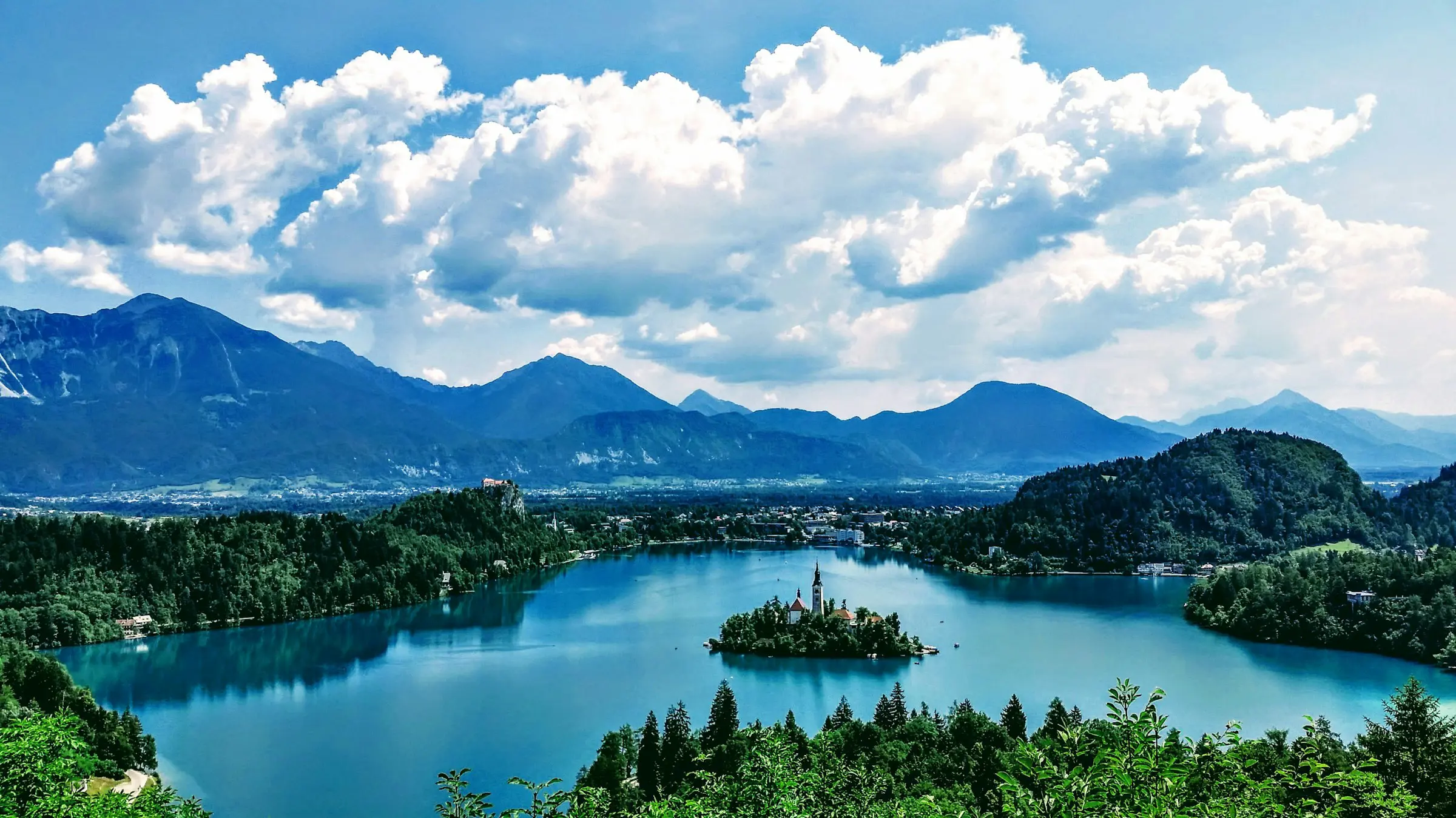 Panorama vy över en sjö med skog och natur kring sig och staden Bled och mäktiga berg i bakgrunden i Slovenien