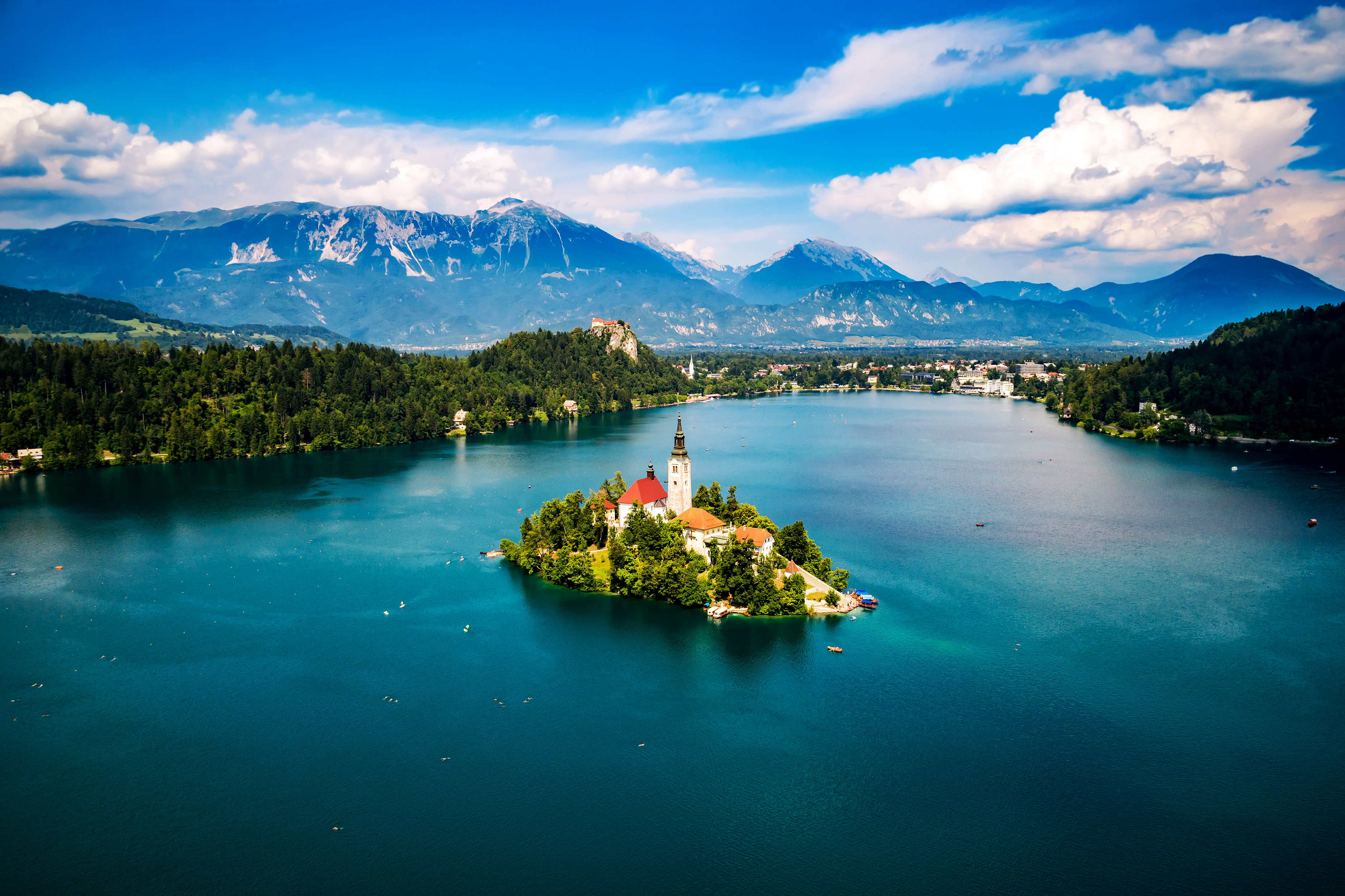 Scenic view of Lake Bled with an island church, surrounded by clear blue waters and lush greenery, set against a backdrop of the Julian Alps under a partly cloudy sky