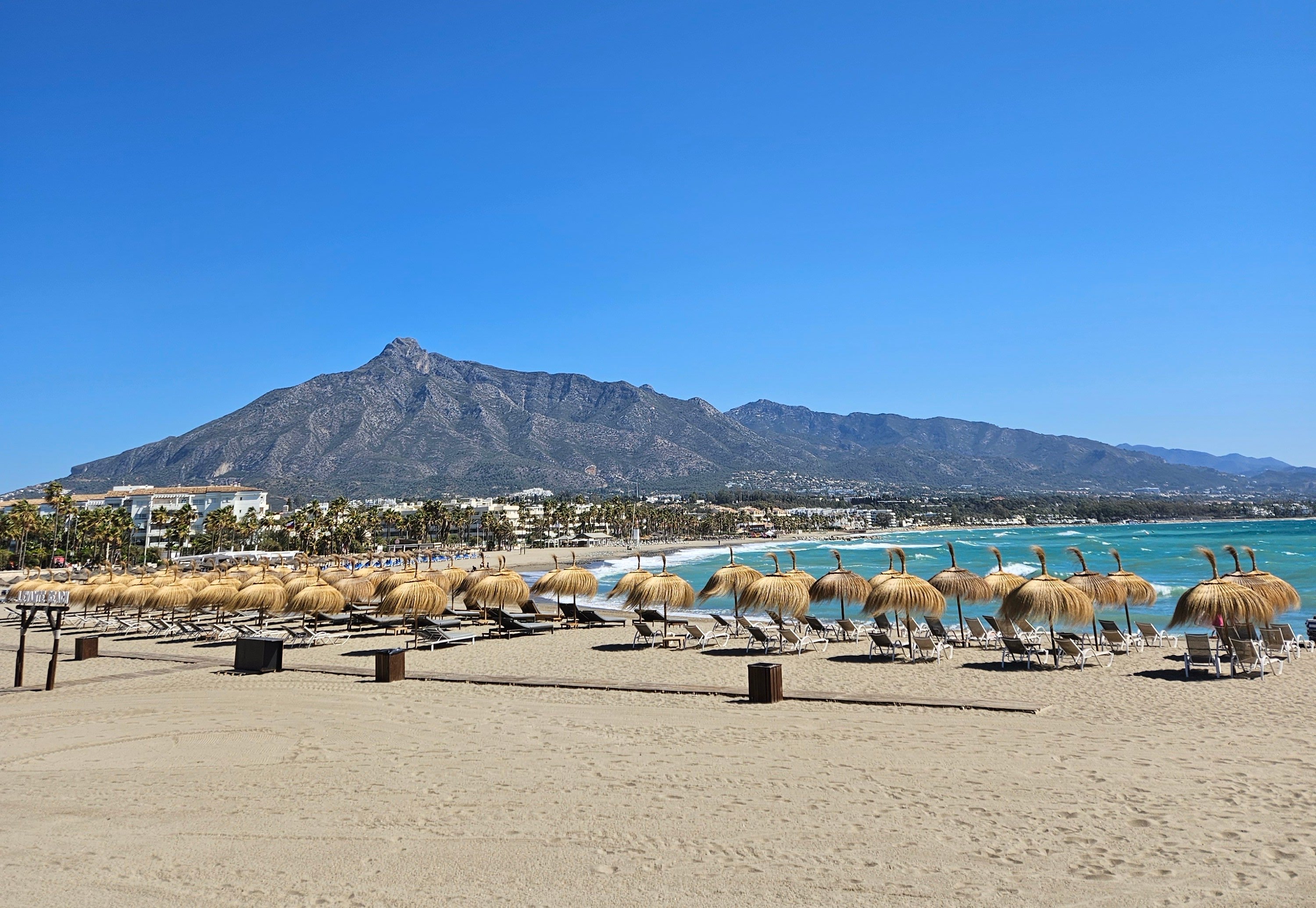 Stor strand med solstolar som möts av blått hav med Marbella stad i bakgrunden 