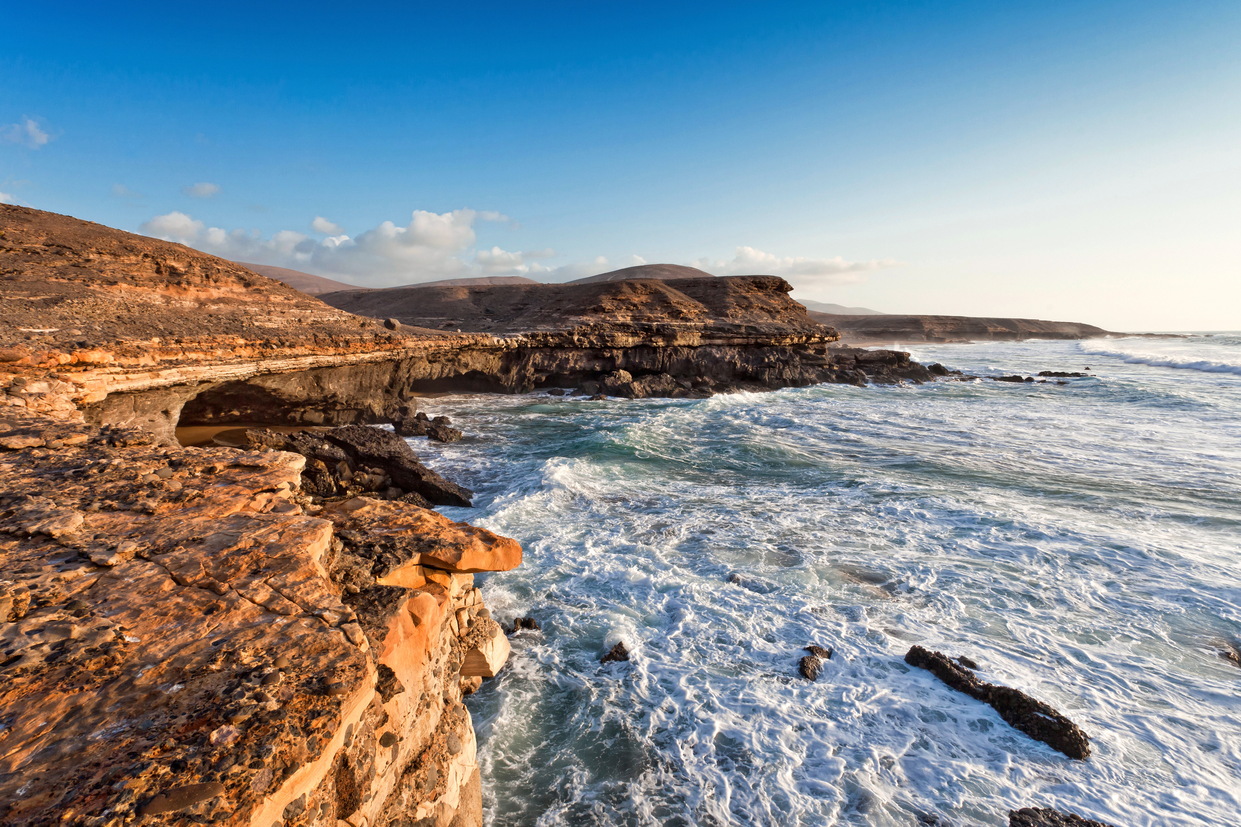 Travel to Fuerteventura - Coastal landscape with rocky formations and turquoise waves at sunset.