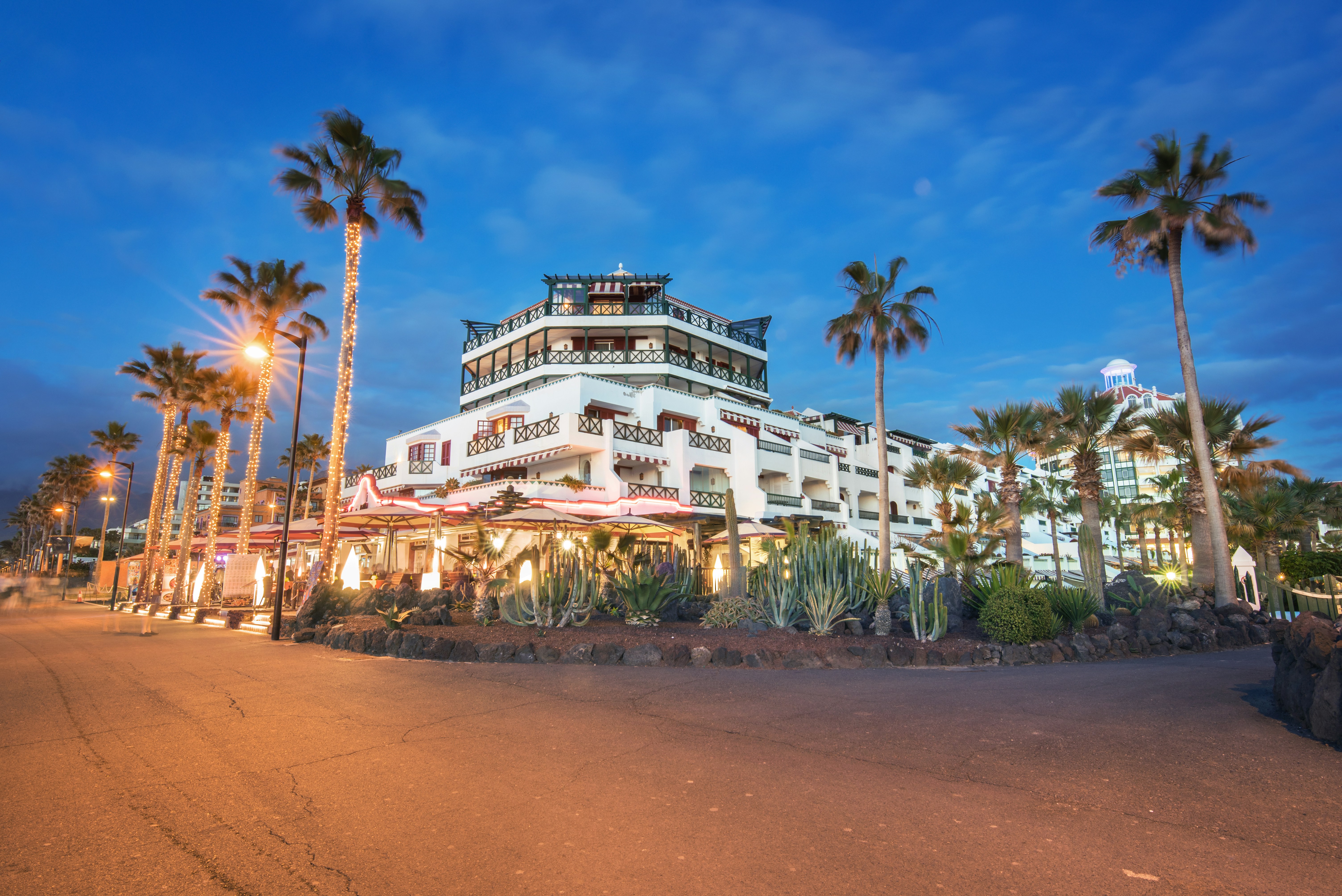 Hotel exterior with palm trees and illuminated facade at dusk, set against a vibrant blue sky