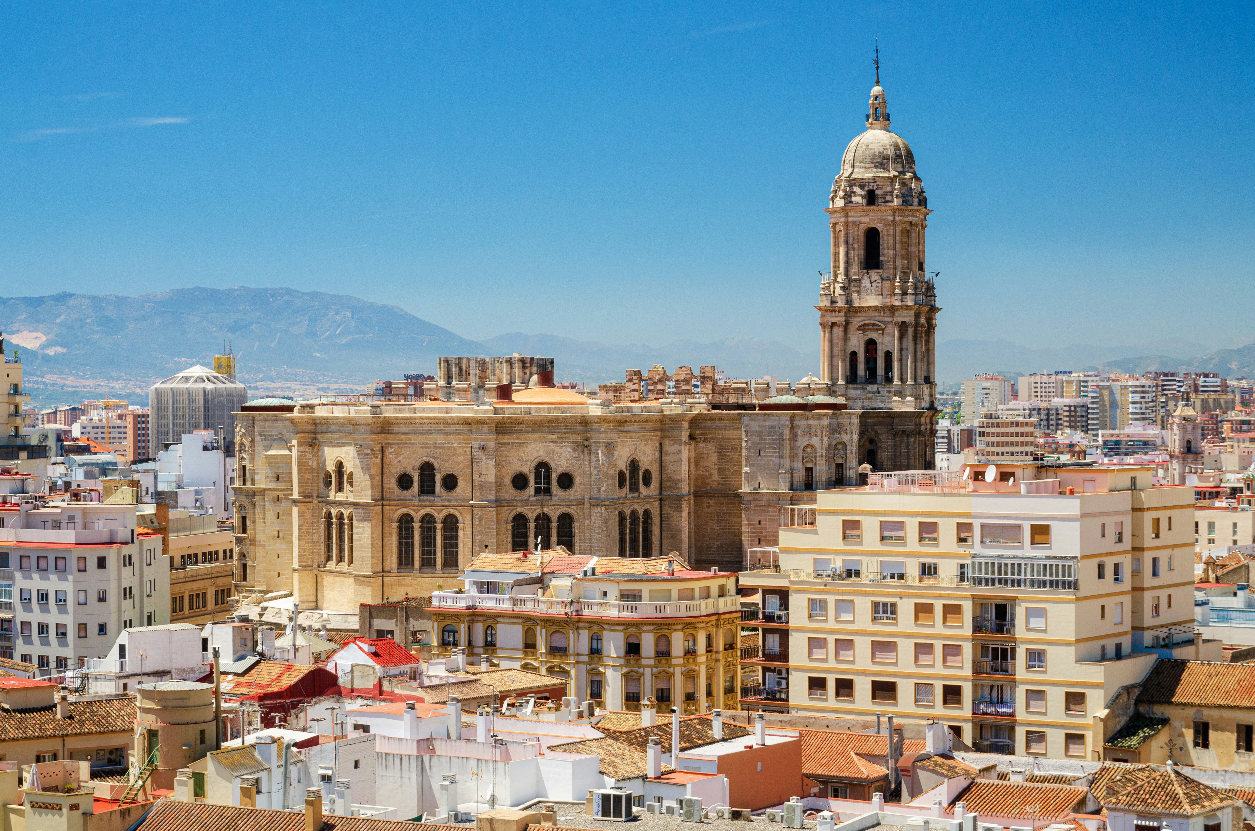 Panoramautsikt över en historisk katedral och omgivande byggnader i Malaga, med berg i bakgrunden och klarblå himmel."