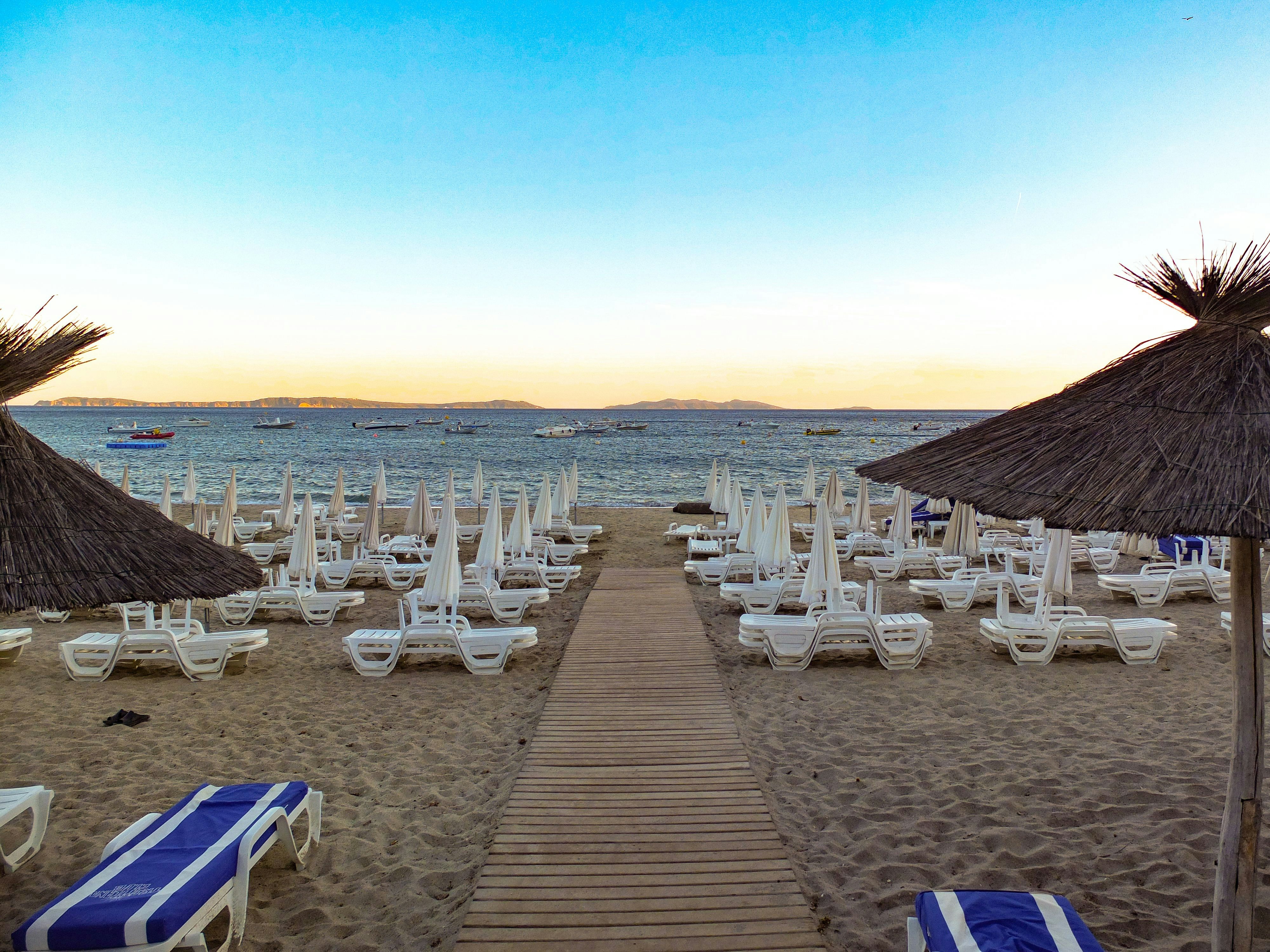 Sandy beach with rows of empty lounge chairs and umbrellas, overlooking a calm sea with several boats near the horizon under a clear blue sky