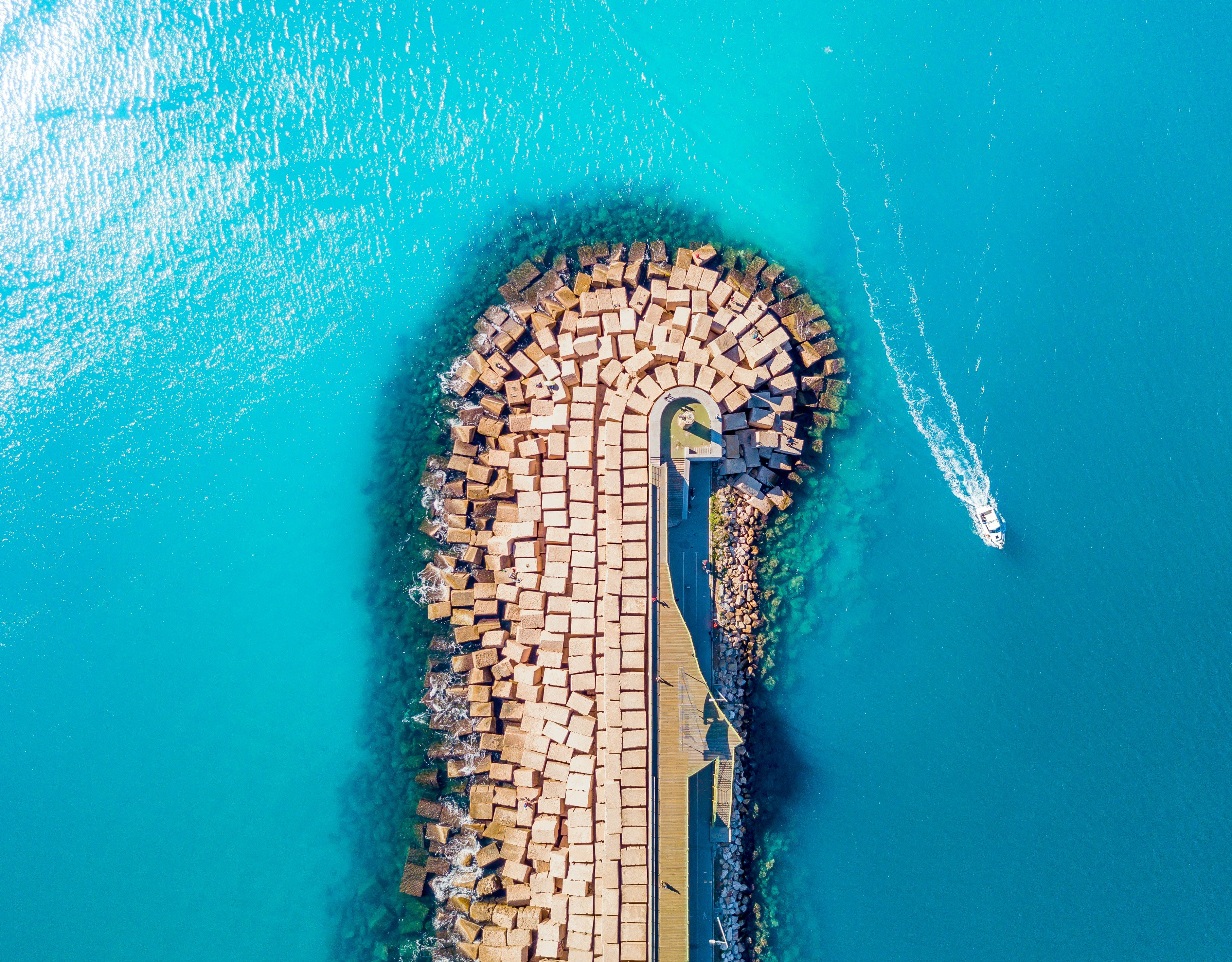 Aerial view of a pier surrounded by turquoise ocean water with a small boat creating a wake, featuring geometric stone formations