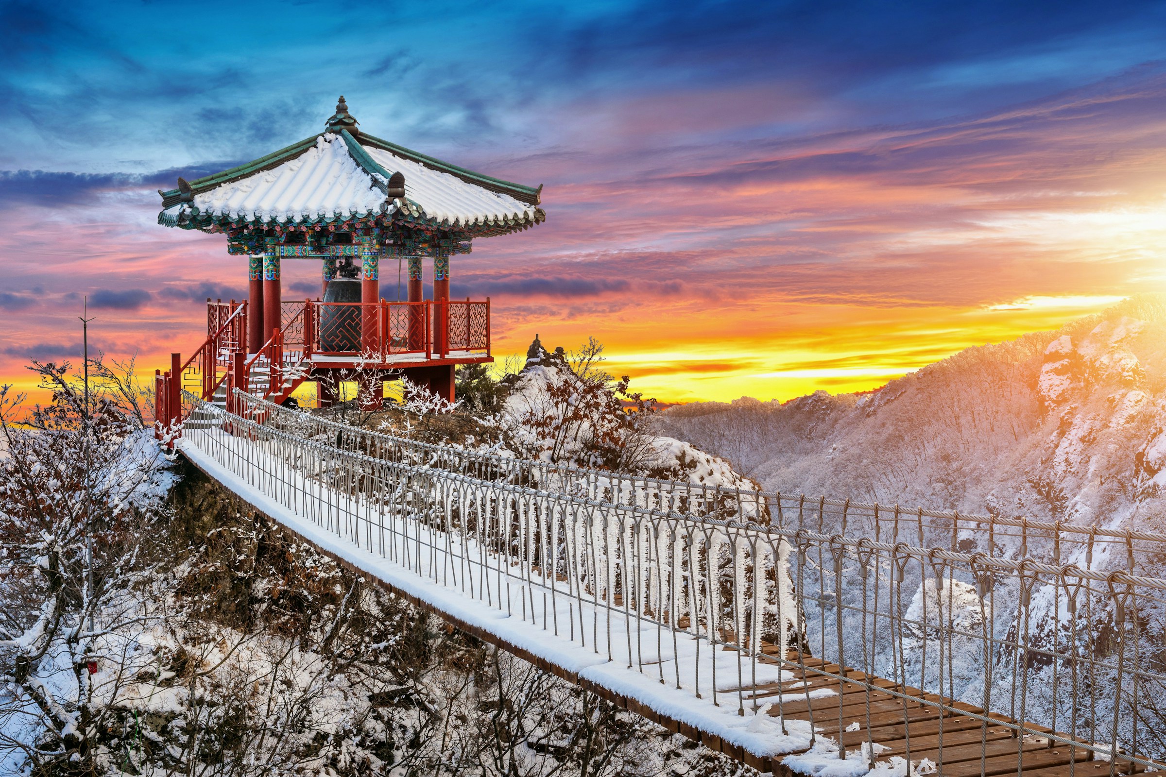 Traditional Asian pavilion on a snowy mountain with a wooden bridge at sunset, featuring vibrant skies and scenic landscape.