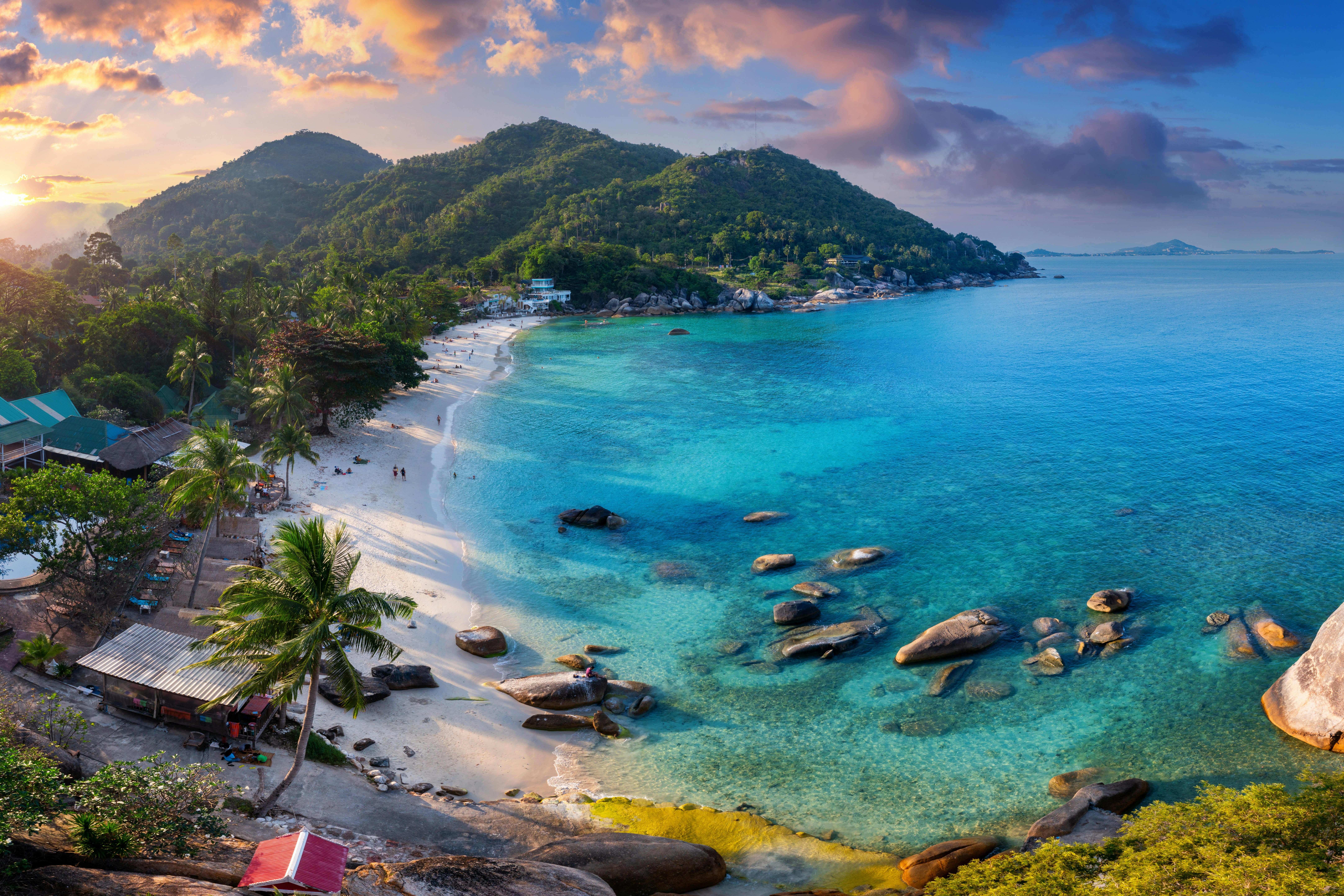 Tropical beach at sunset with turquoise water and lush green hills, featuring palm trees and scattered rocks along the shore