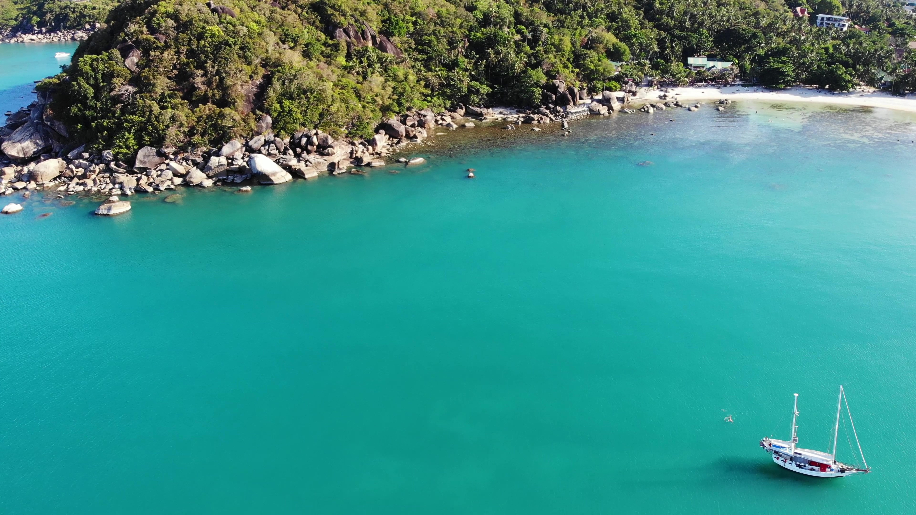 Aerial view of a sailboat in turquoise water near a lush, rocky island coastline with a sandy beach