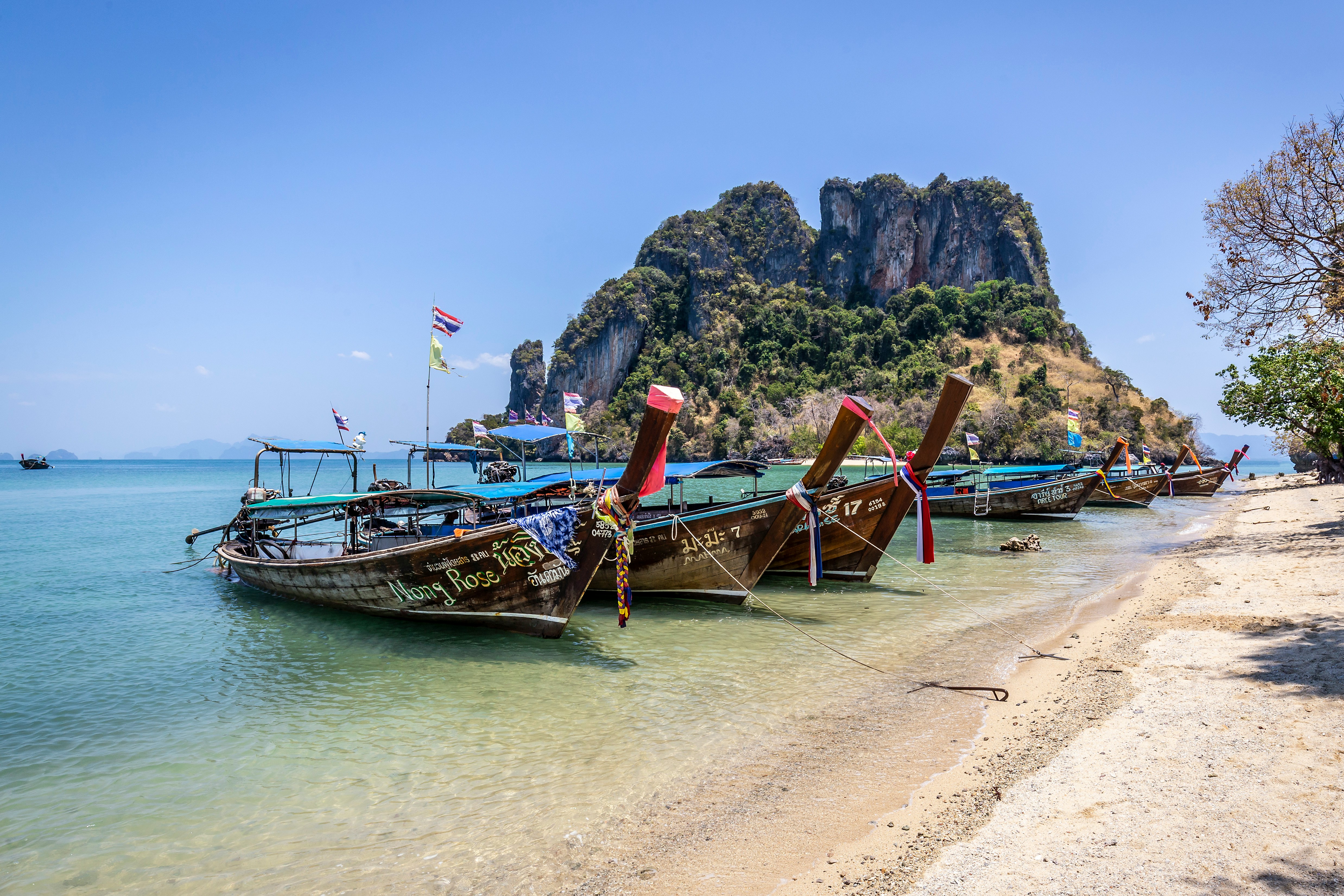 Traditional long-tail boats anchored on a sandy beach in Thailand with a picturesque limestone cliff in the background.