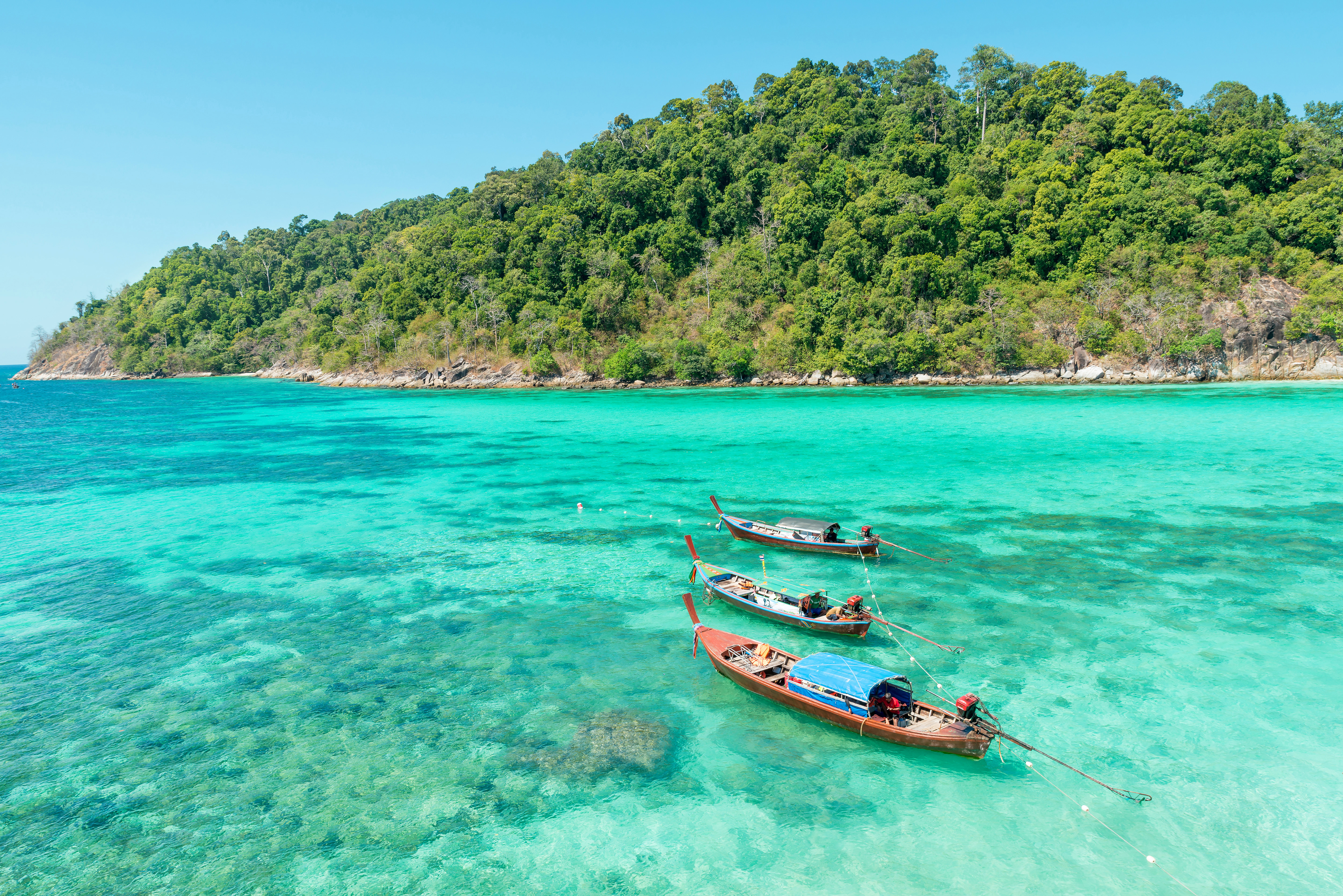 Three traditional longtail boats floating on clear turquoise waters near a lush green island under a bright blue sky
