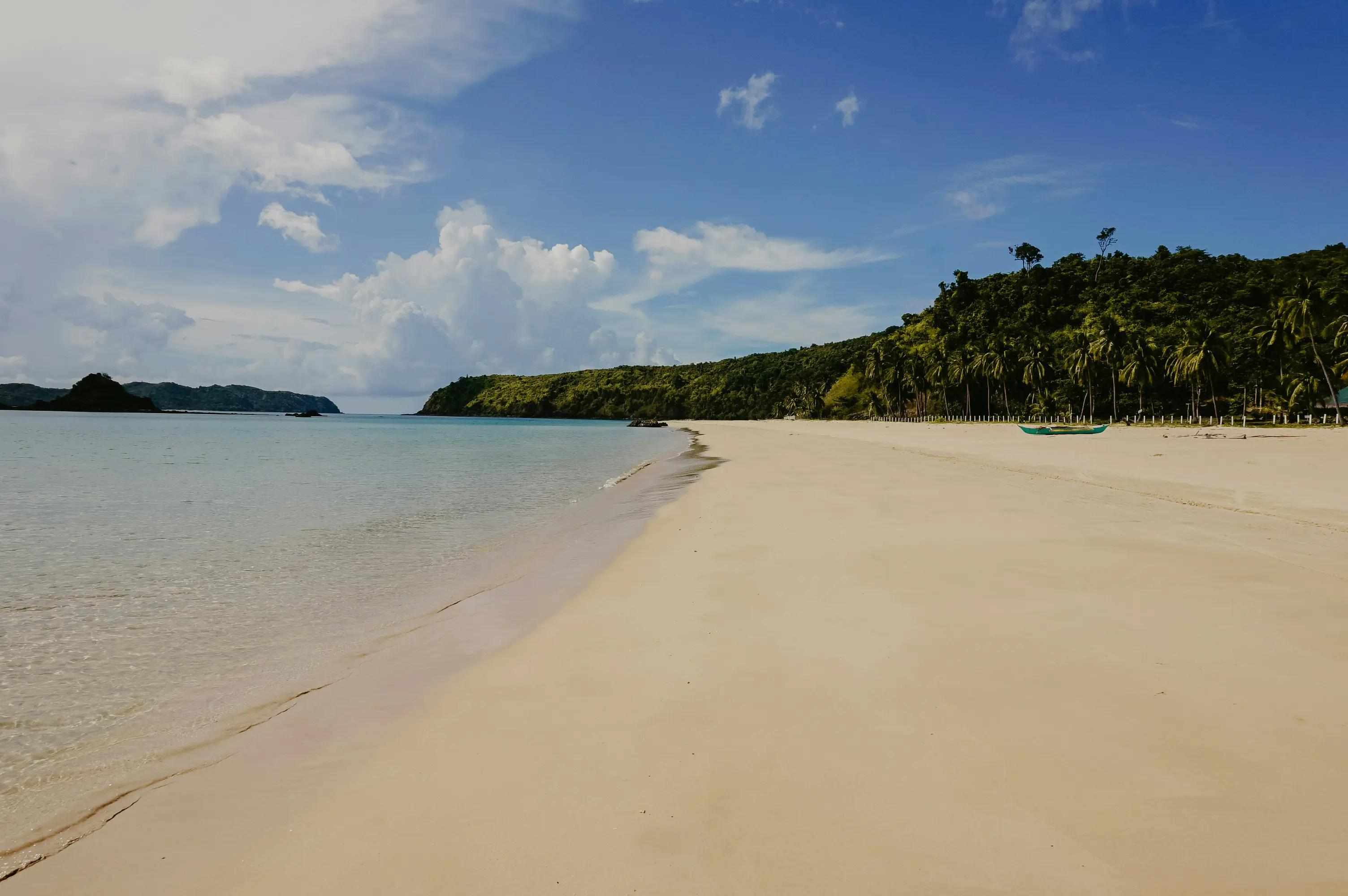 Sandy beach with clear blue water and tree-covered hills under a sunny sky
