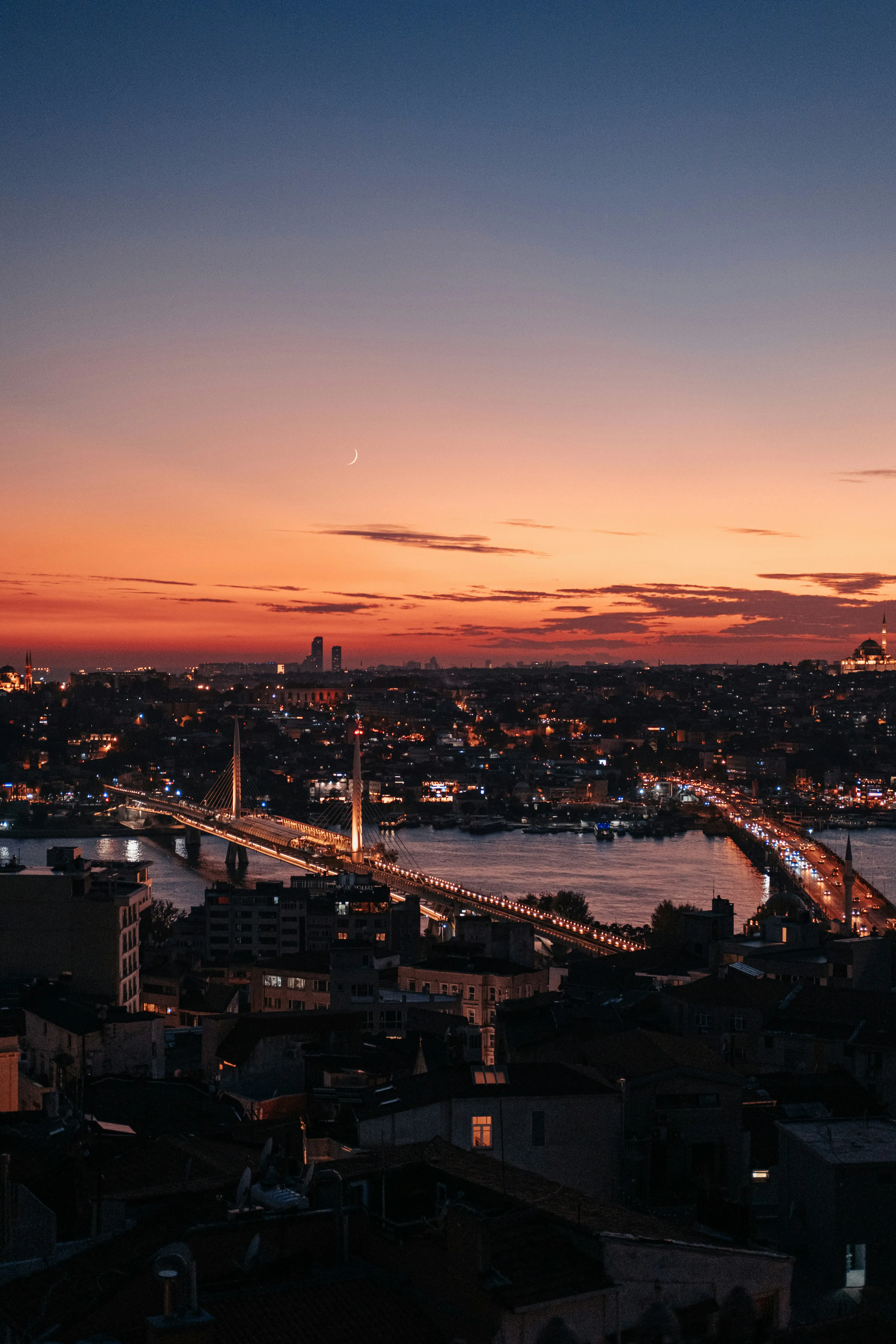 Cityscape of Istanbul at sunset featuring a lit bridge and crescent moon in the colorful sky