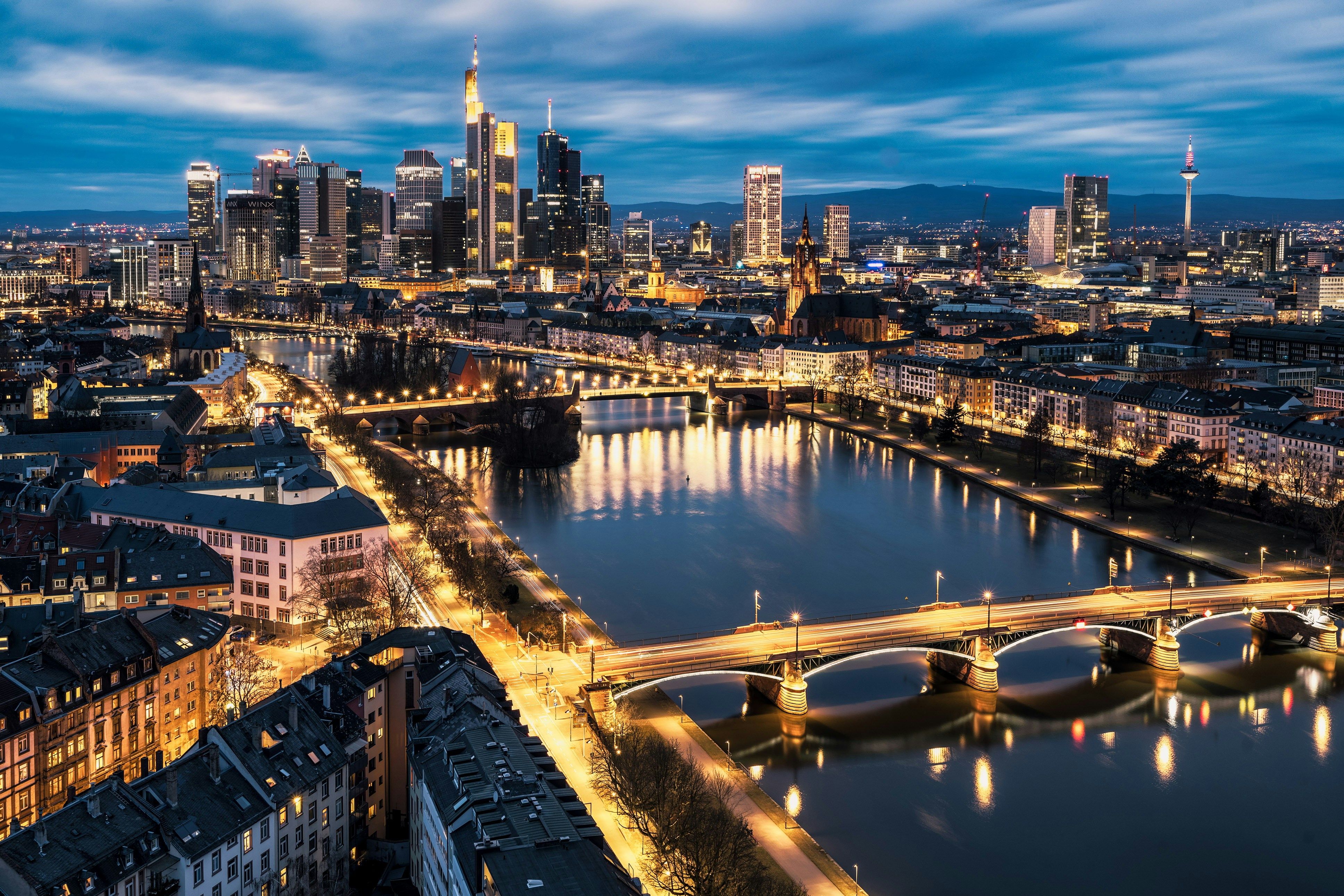 Skyline of Frankfurt, Germany at dusk with illuminated skyscrapers, reflecting on the Main River and visible bridges, under a blue, cloudy sky
