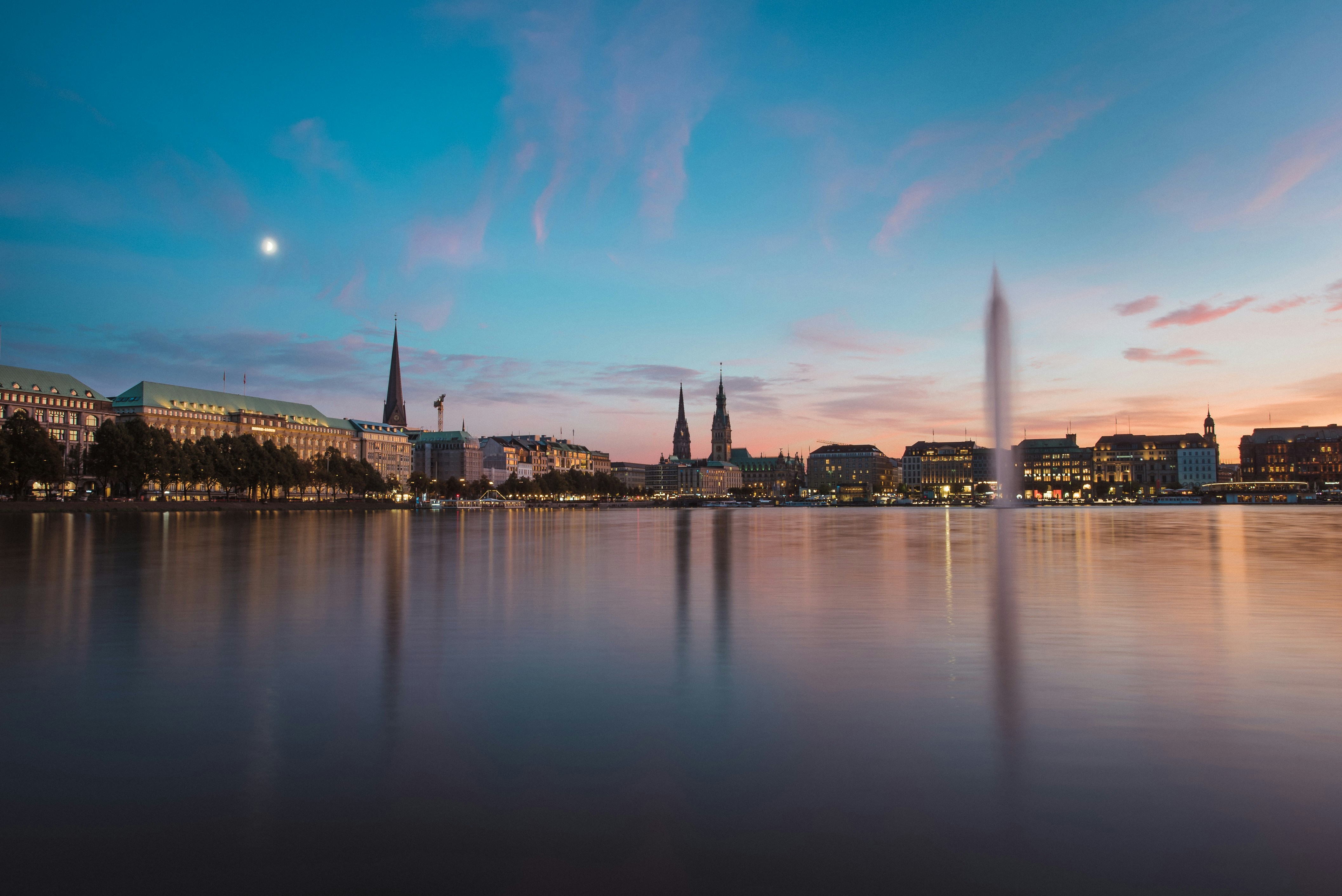 Twilight view of Hamburg cityscape reflected in Alster Lake, featuring buildings and church spires against a colorful evening sky.