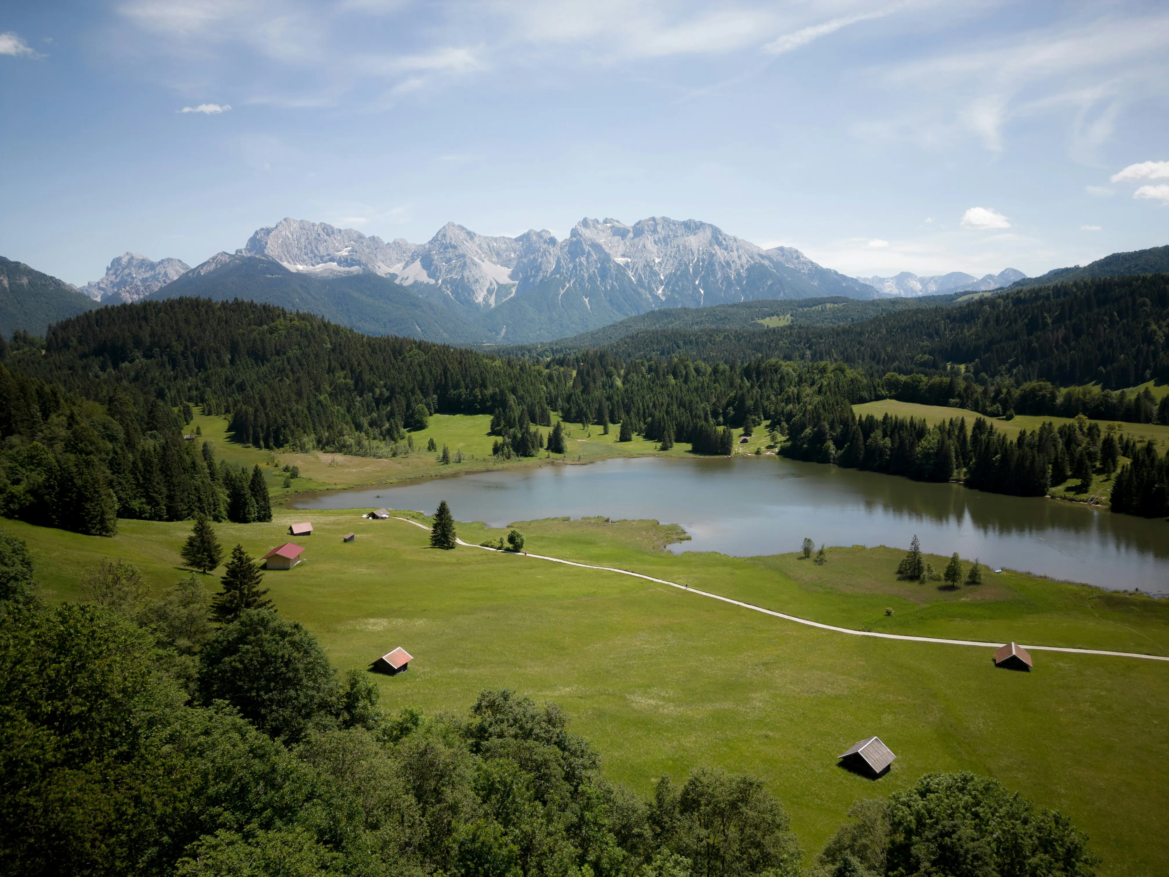 Mountain landscape with a serene lake surrounded by green meadows and forest, with cabins and distant snow-capped peaks under a blue sky