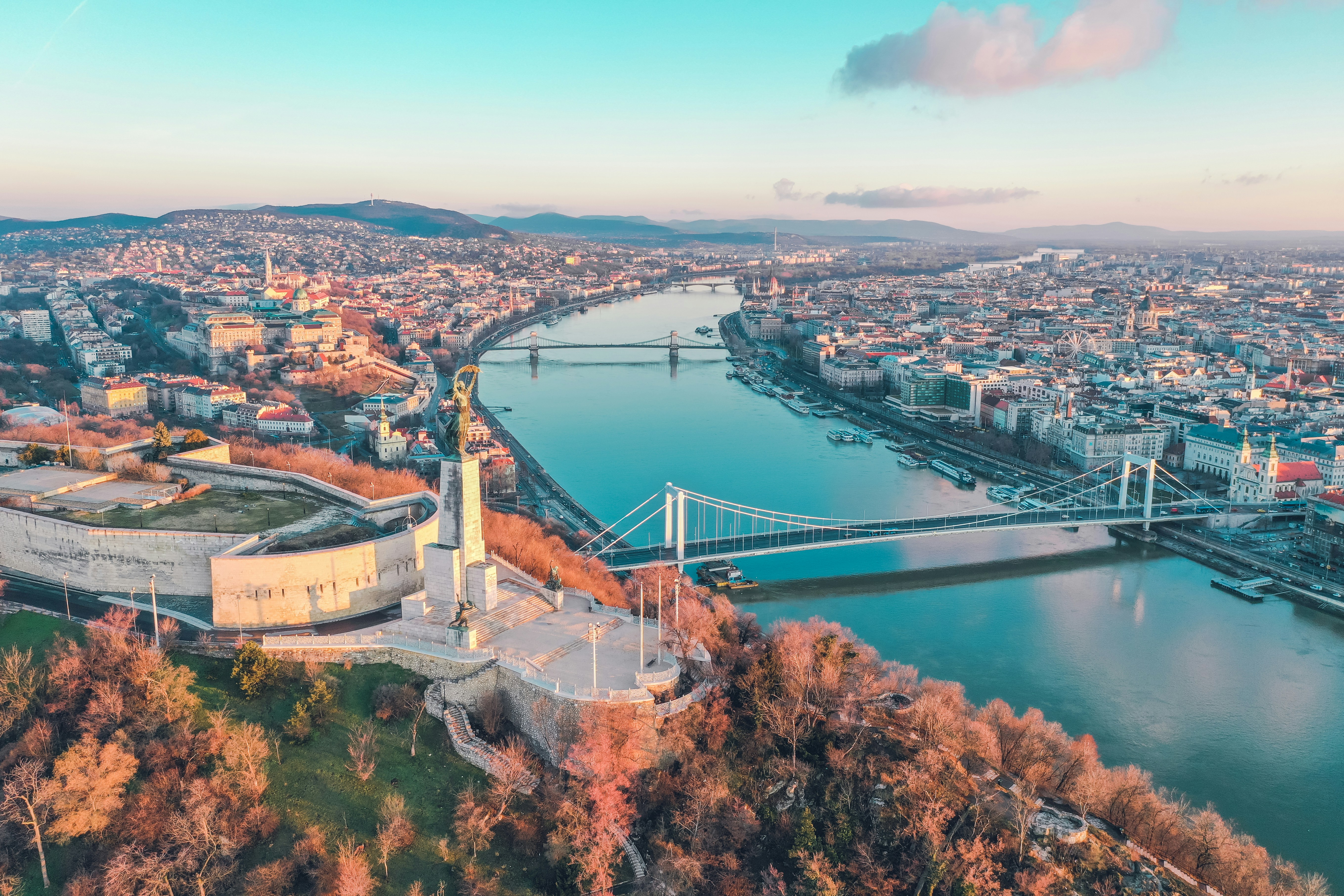 Aerial view of Budapest, Hungary, featuring the Danube River, iconic Chain Bridge, and historic architecture with Gellért Hill and Liberty Statue in the foreground during sunset