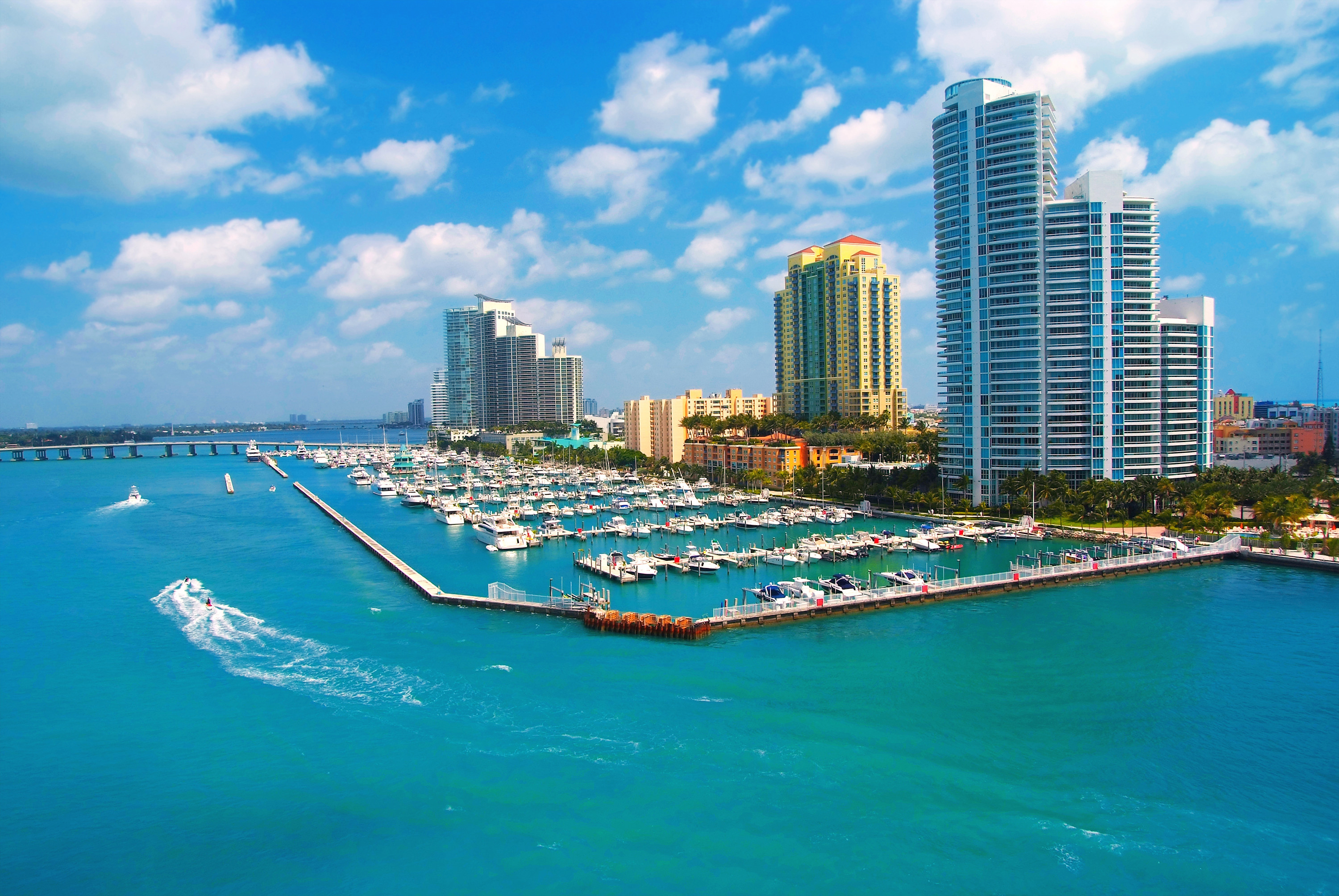 Aerial view of Miami Beach Marina with luxury yachts, turquoise water, and modern high-rise buildings under a bright blue sky with scattered clouds