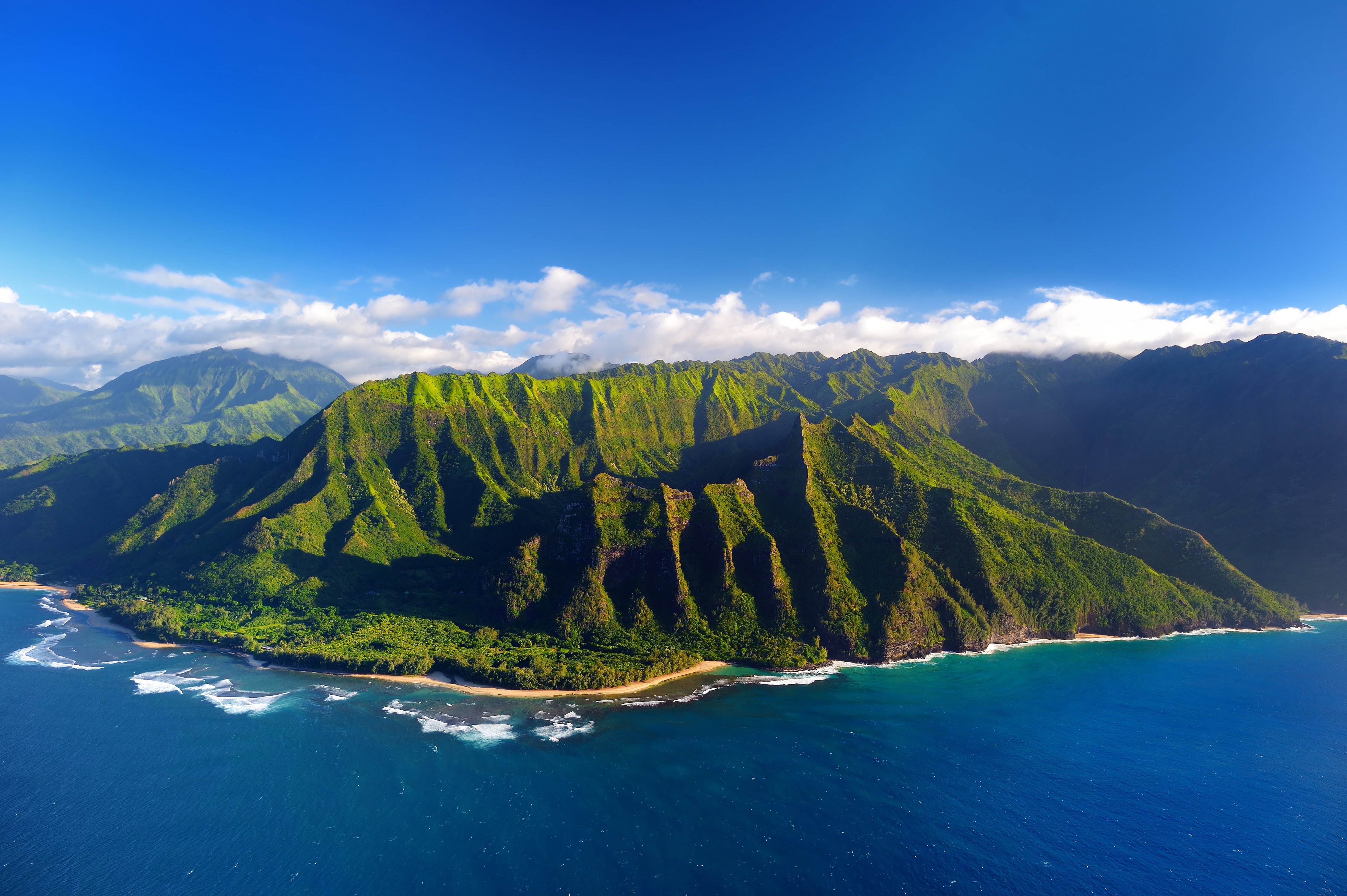 Aerial view of the lush, green cliffs of the Na Pali Coast on Kauai Island, Hawaii, with the Pacific Ocean in the foreground under a clear blue sky