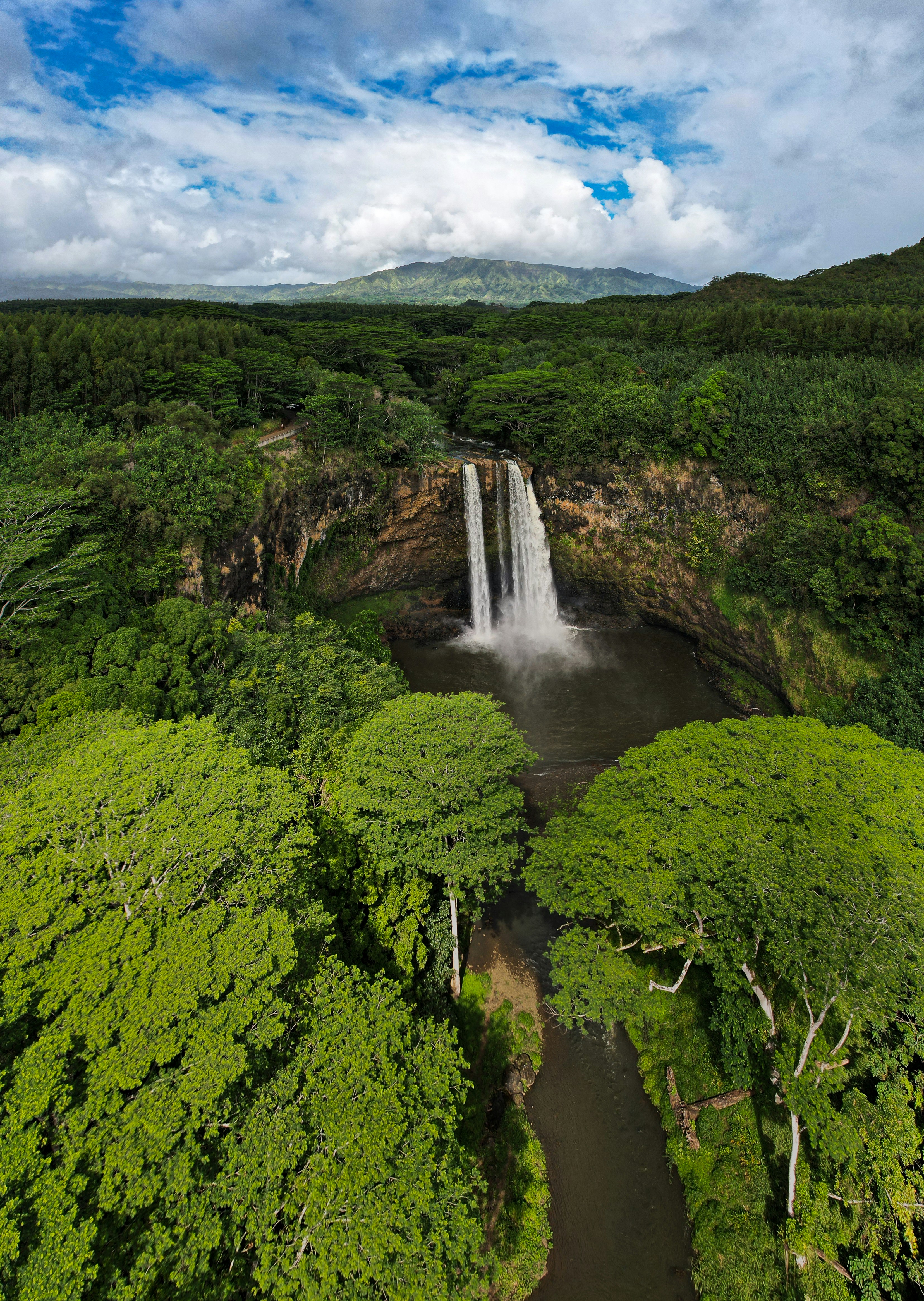 Aerial view of a waterfall surrounded by lush green forest, with mountains in the background and a partly cloudy sky above
