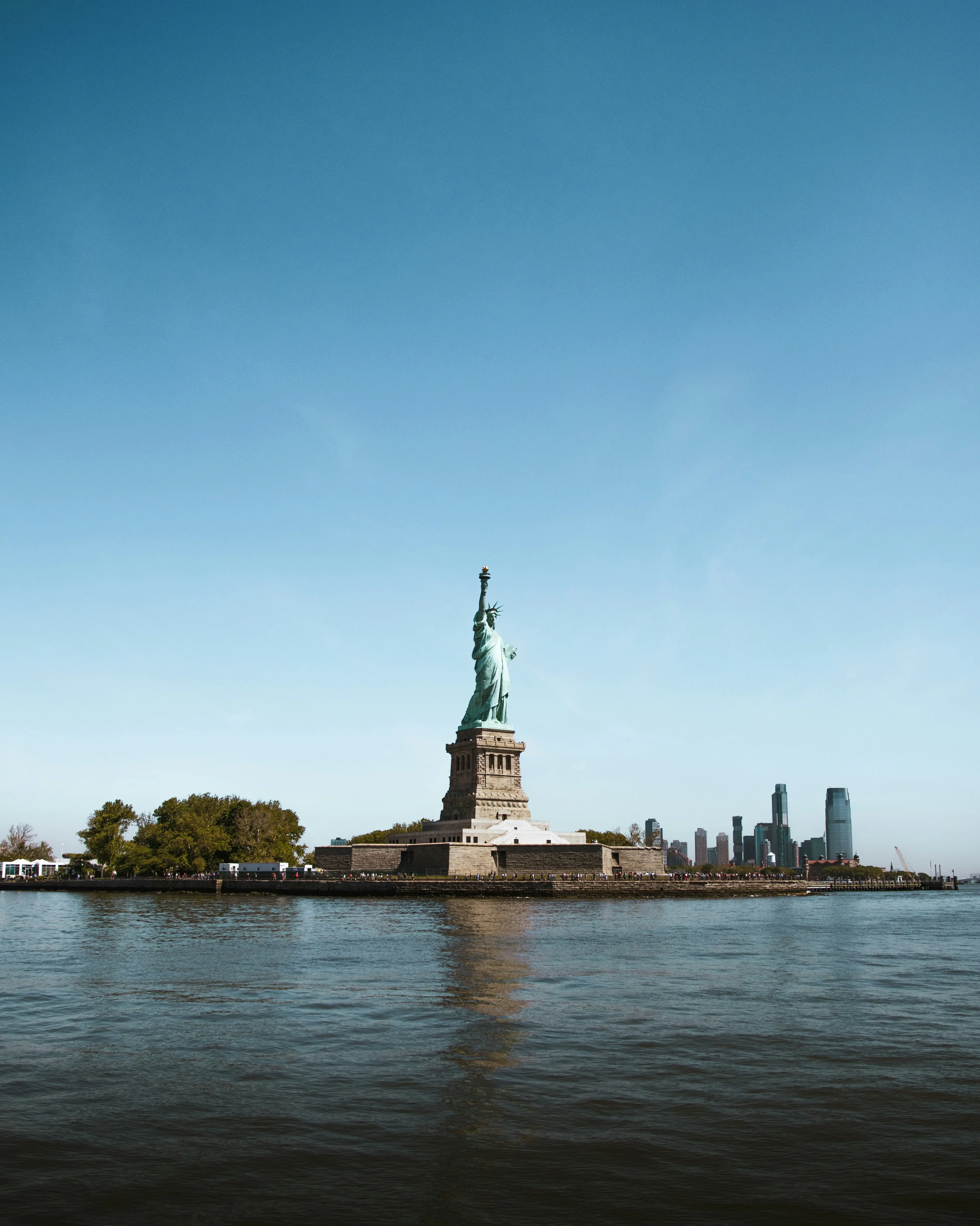 Frihetsgudinnan mot en klarblå himmel, belägen på Liberty Island med New Yorks skyline i bakgrunden